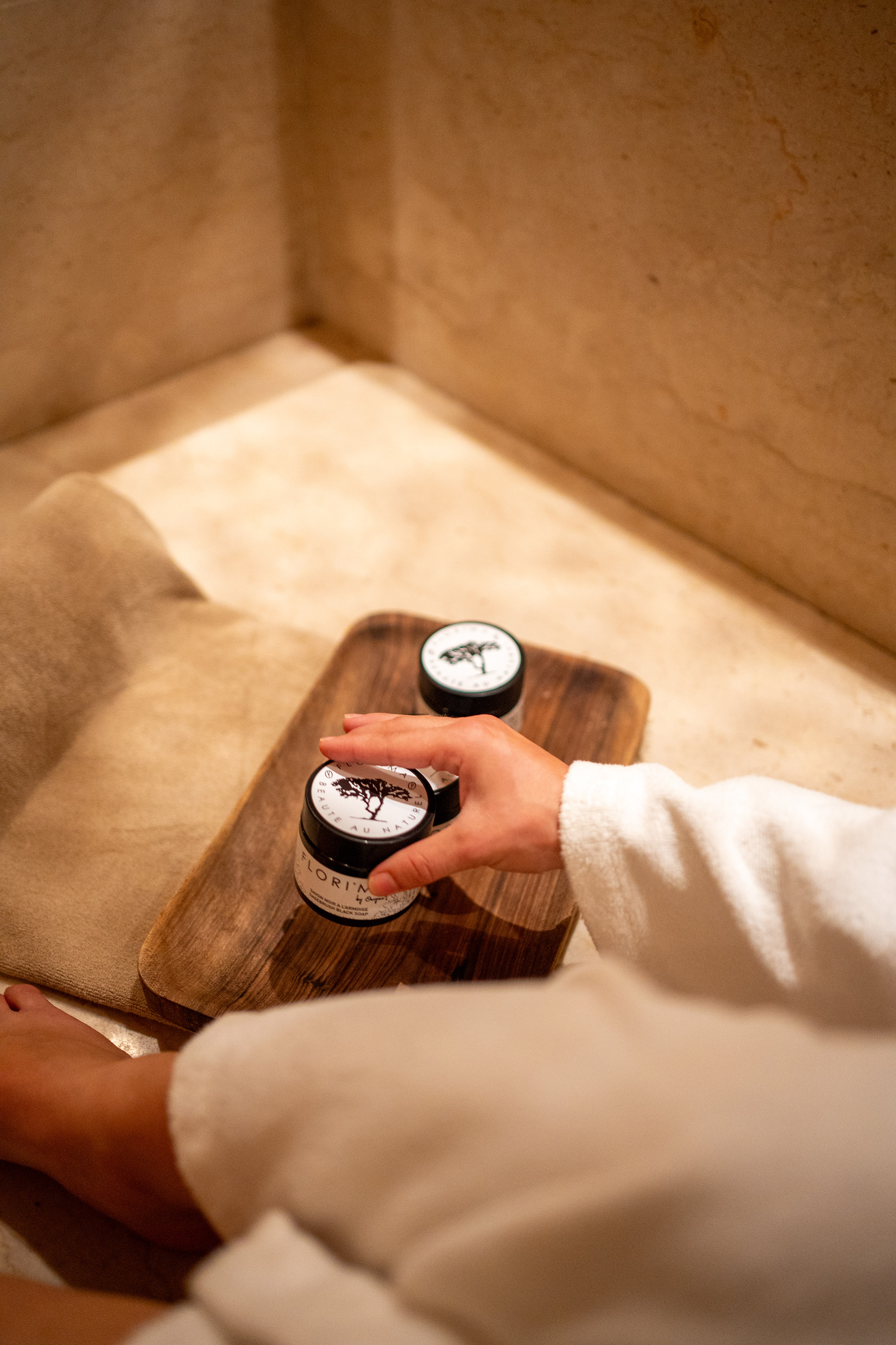 Hand in robe selecting Moroccan black soap jar from a wooden tray. Spa beauty.