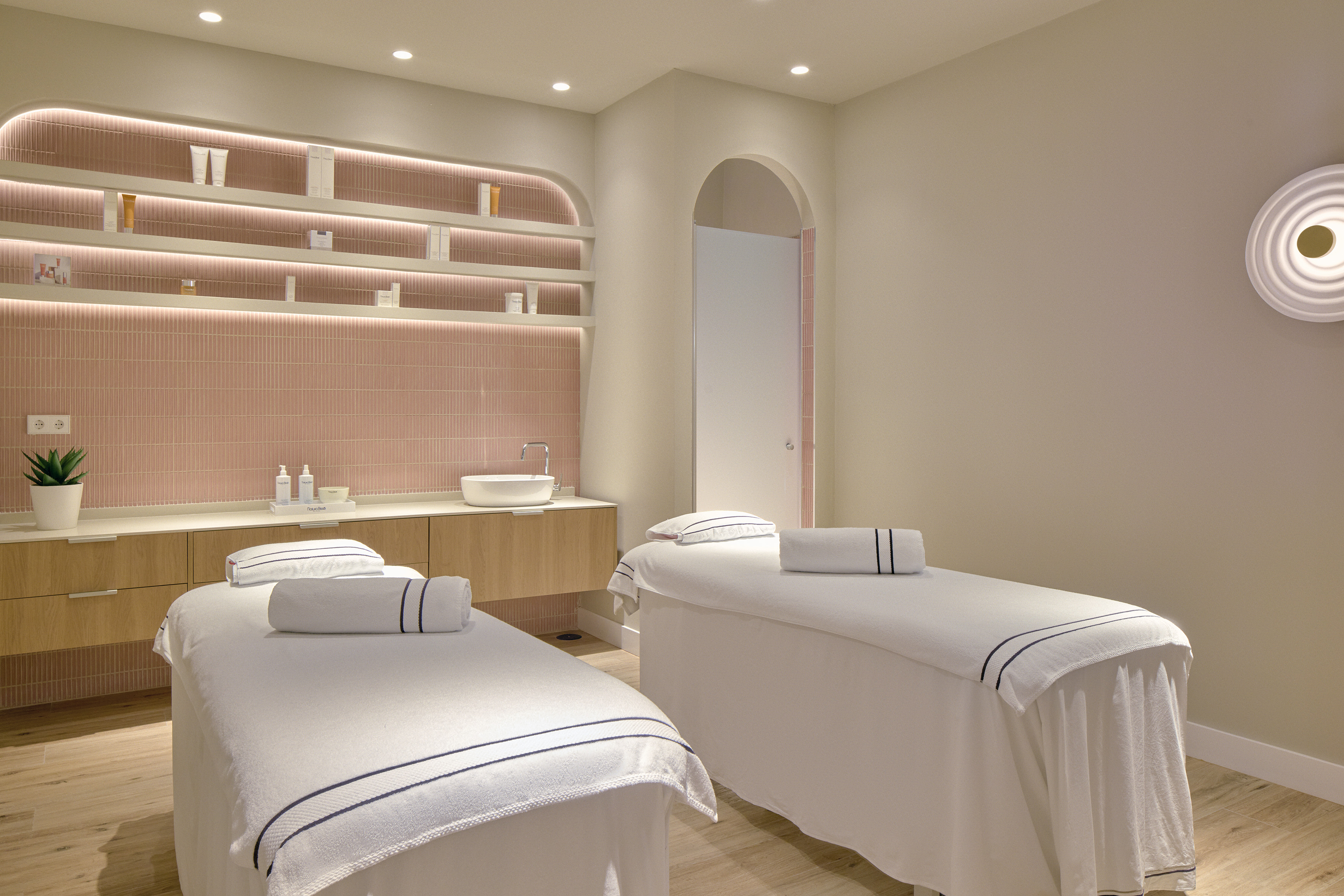 Bright spa treatment room with two massage beds and pink tiled wall.