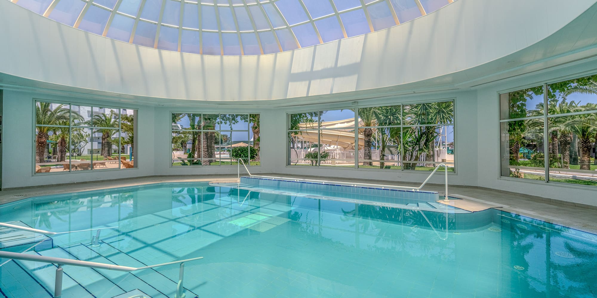 Indoor resort pool with glass dome, palm trees, and visible water slide.