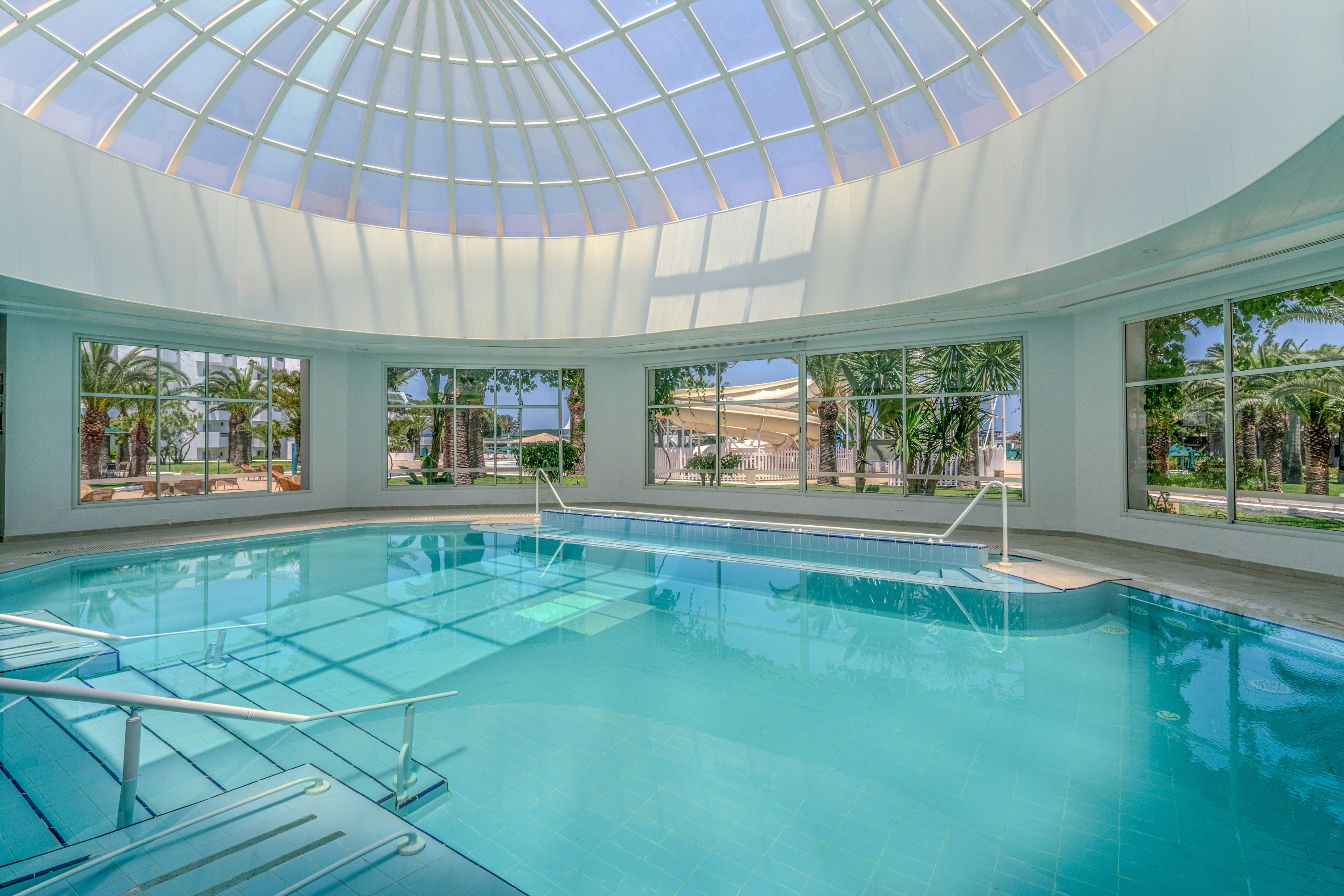 Indoor resort pool with glass dome, palm trees, and visible water slide.