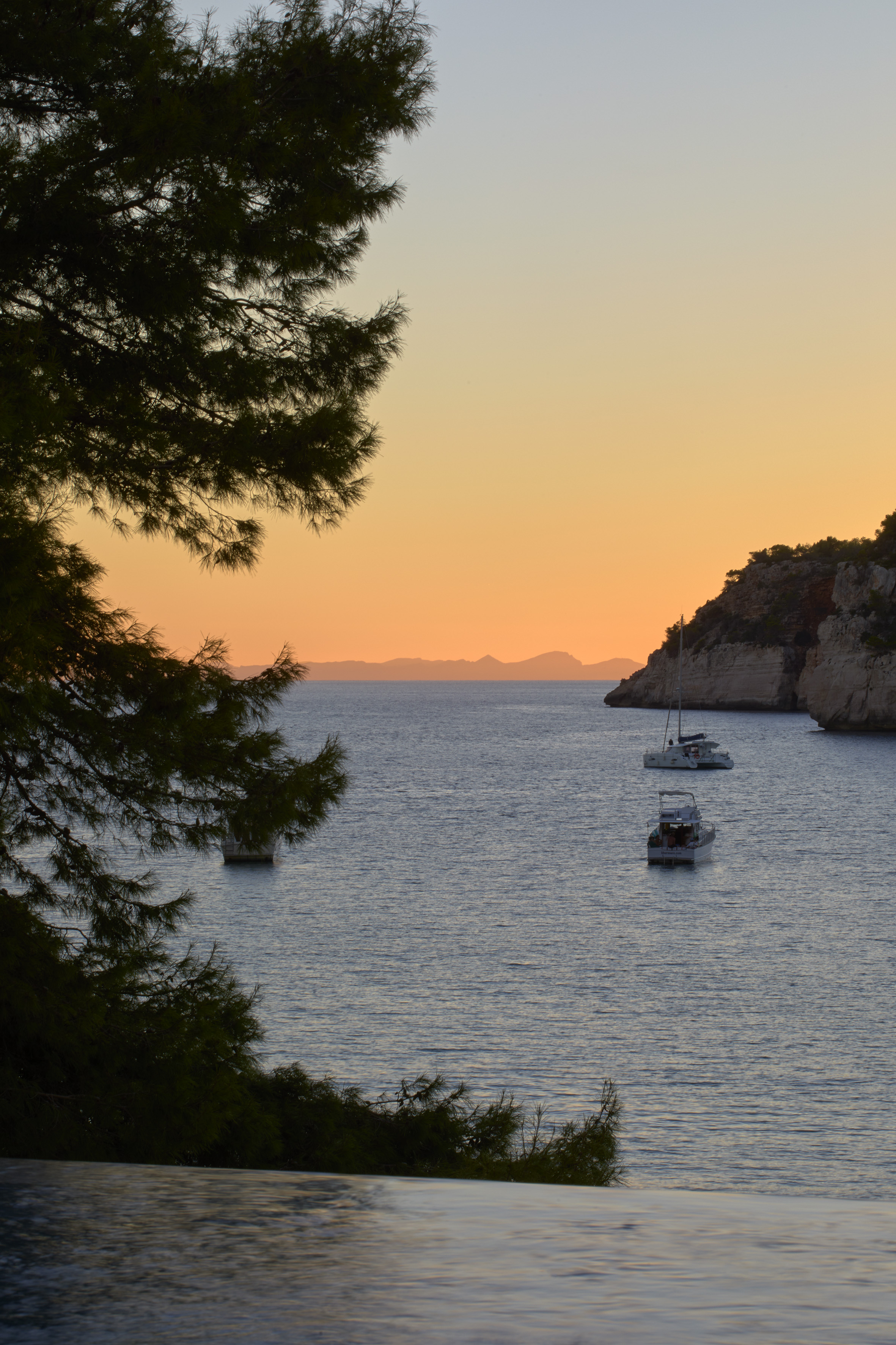 Orange sunset over calm bay with boats, cliffs, and silhouetted pine.
