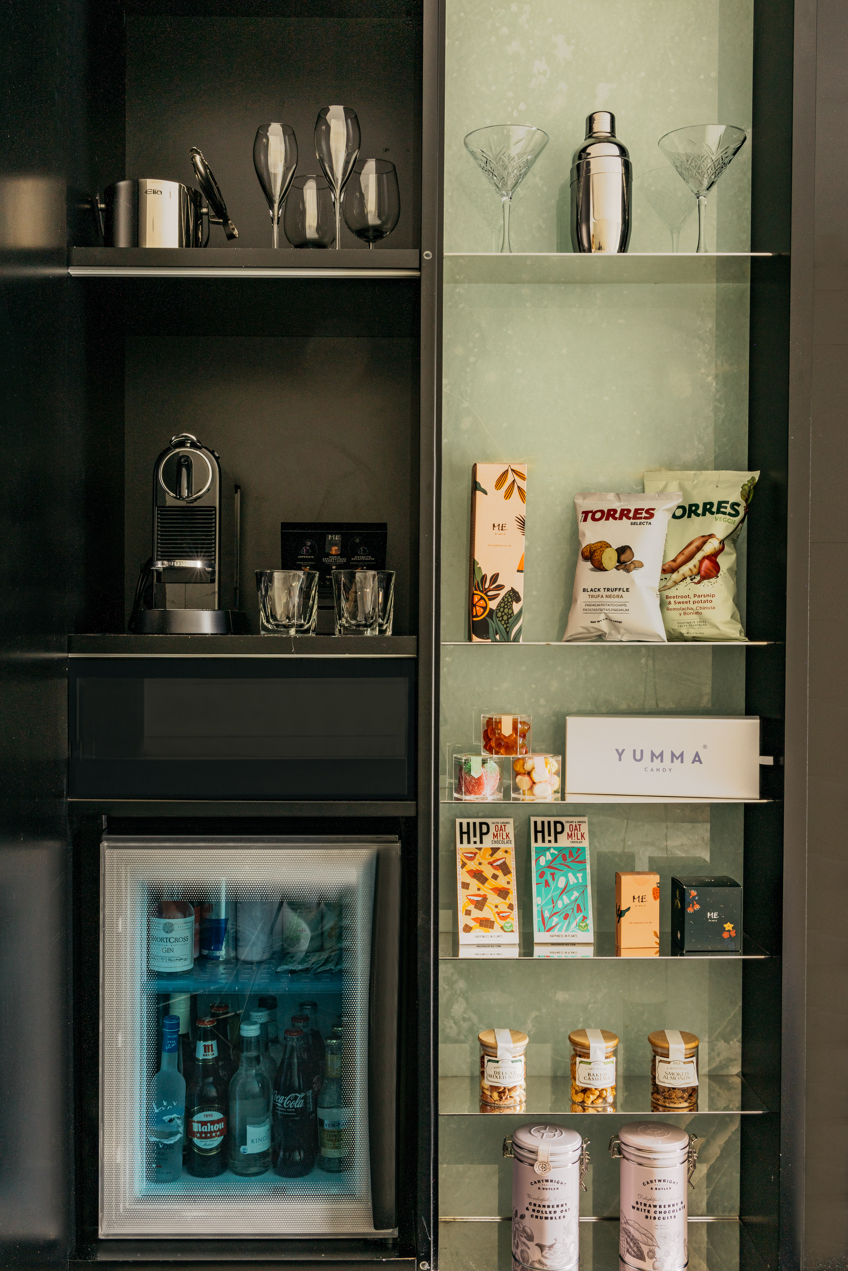 Luxury hotel minibar with Nespresso machine, cocktail set, assorted snacks, and drinks fridge.
