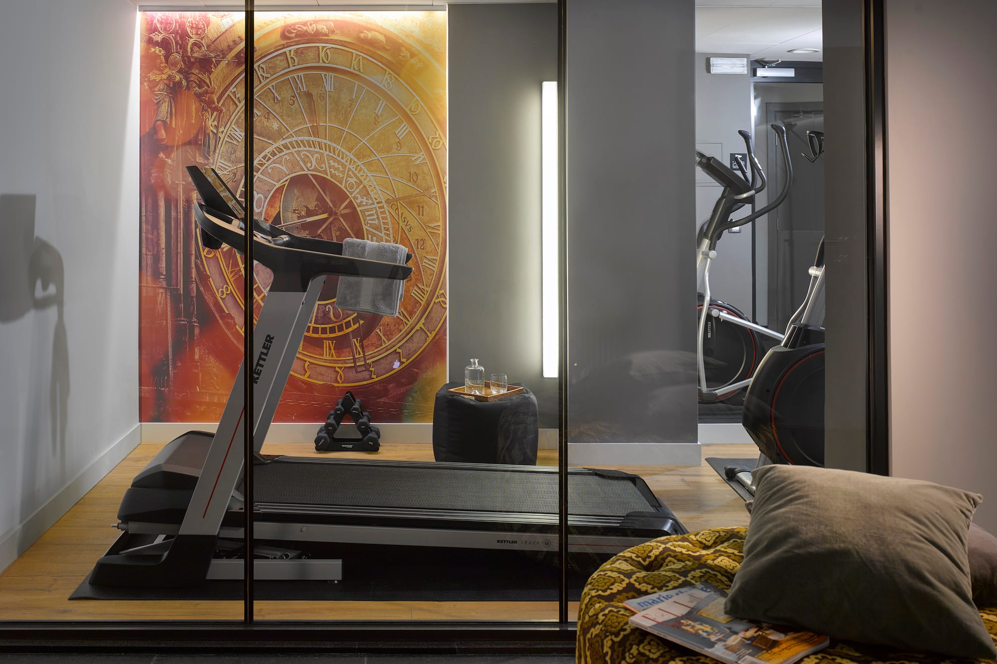 Home gym with treadmill, elliptical, dumbbells, and decorative clock mural.