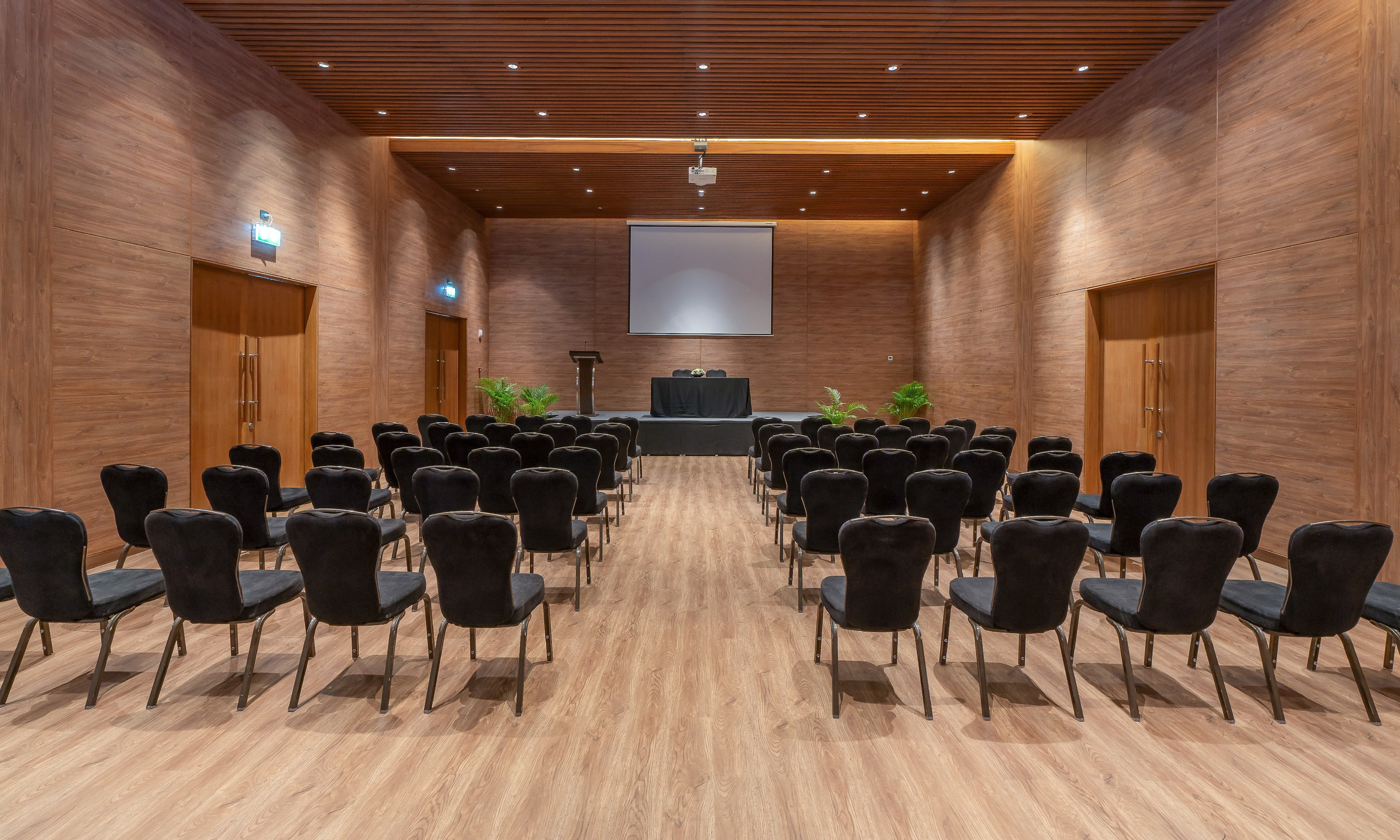 Wood-paneled meeting room with black seating, stage, and projection screen setup.