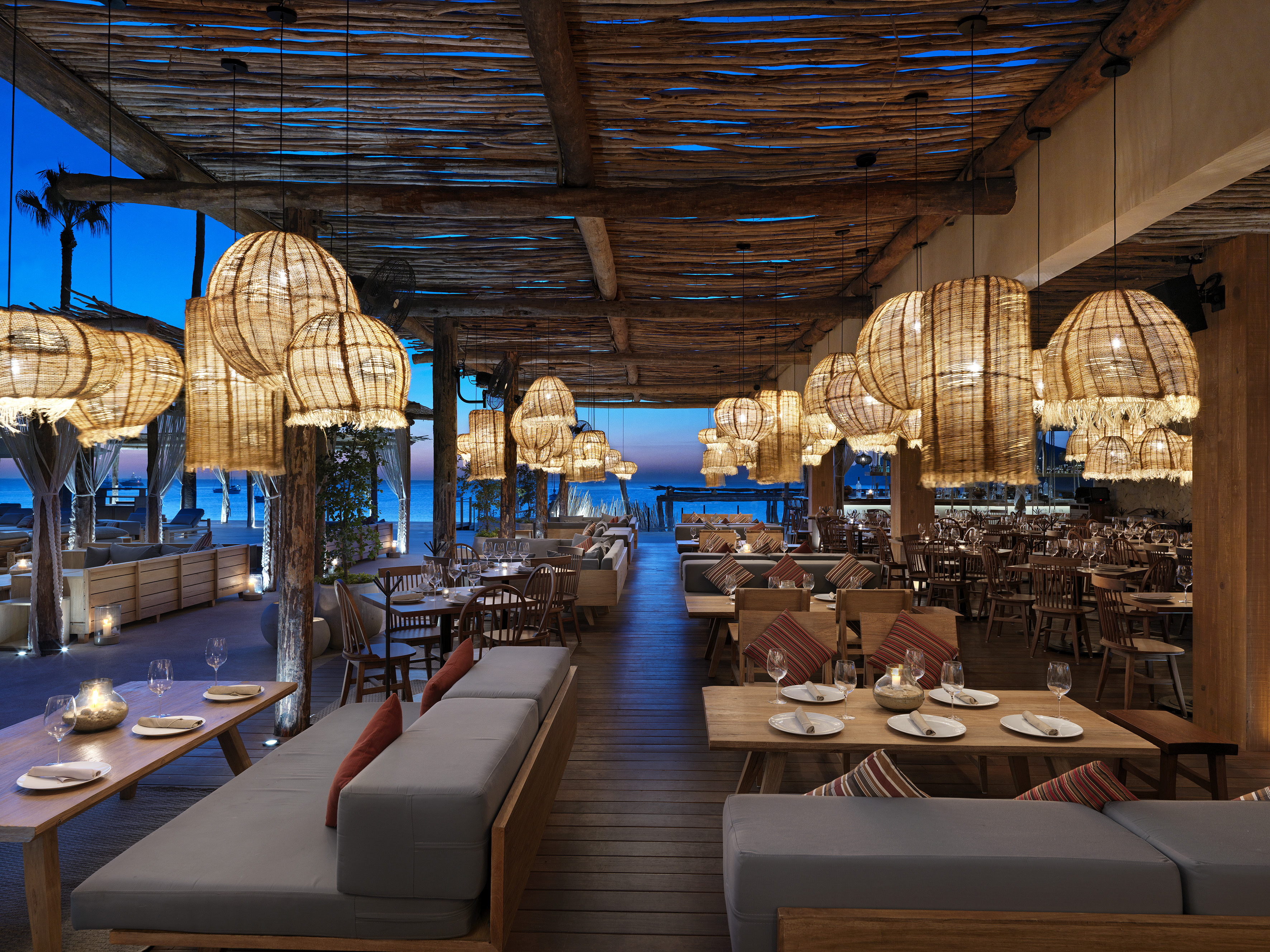 Rustic beachside restaurant with woven pendant lights, wooden decor, and sea view at dusk.