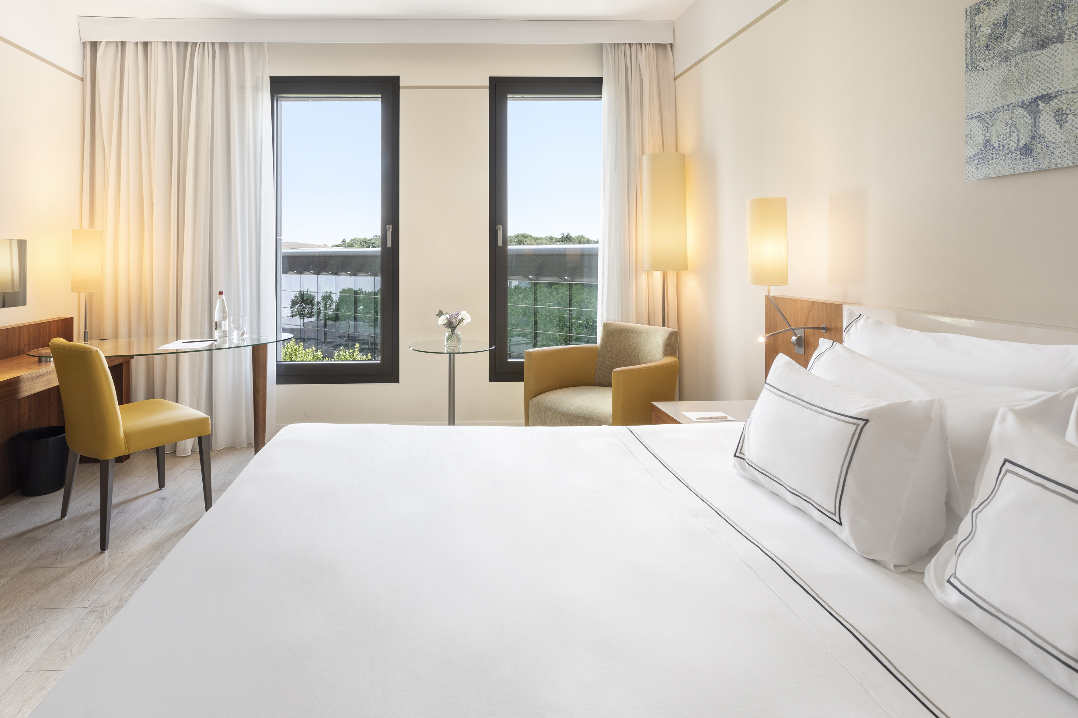 Modern hotel room with white bed, desk, yellow chair, and sunlit windows.