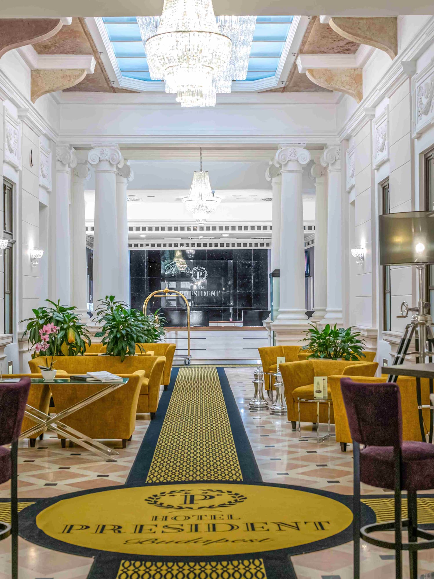 Elegant Hotel President Budapest lobby with chandeliers, columns, and gold seating.
