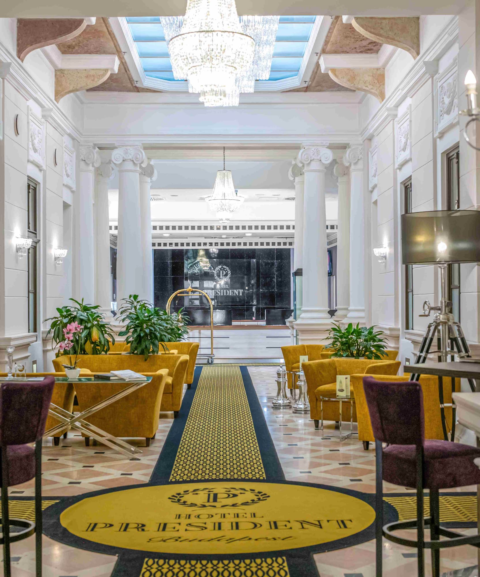 Elegant Hotel President Budapest lobby with chandeliers, columns, and gold seating.