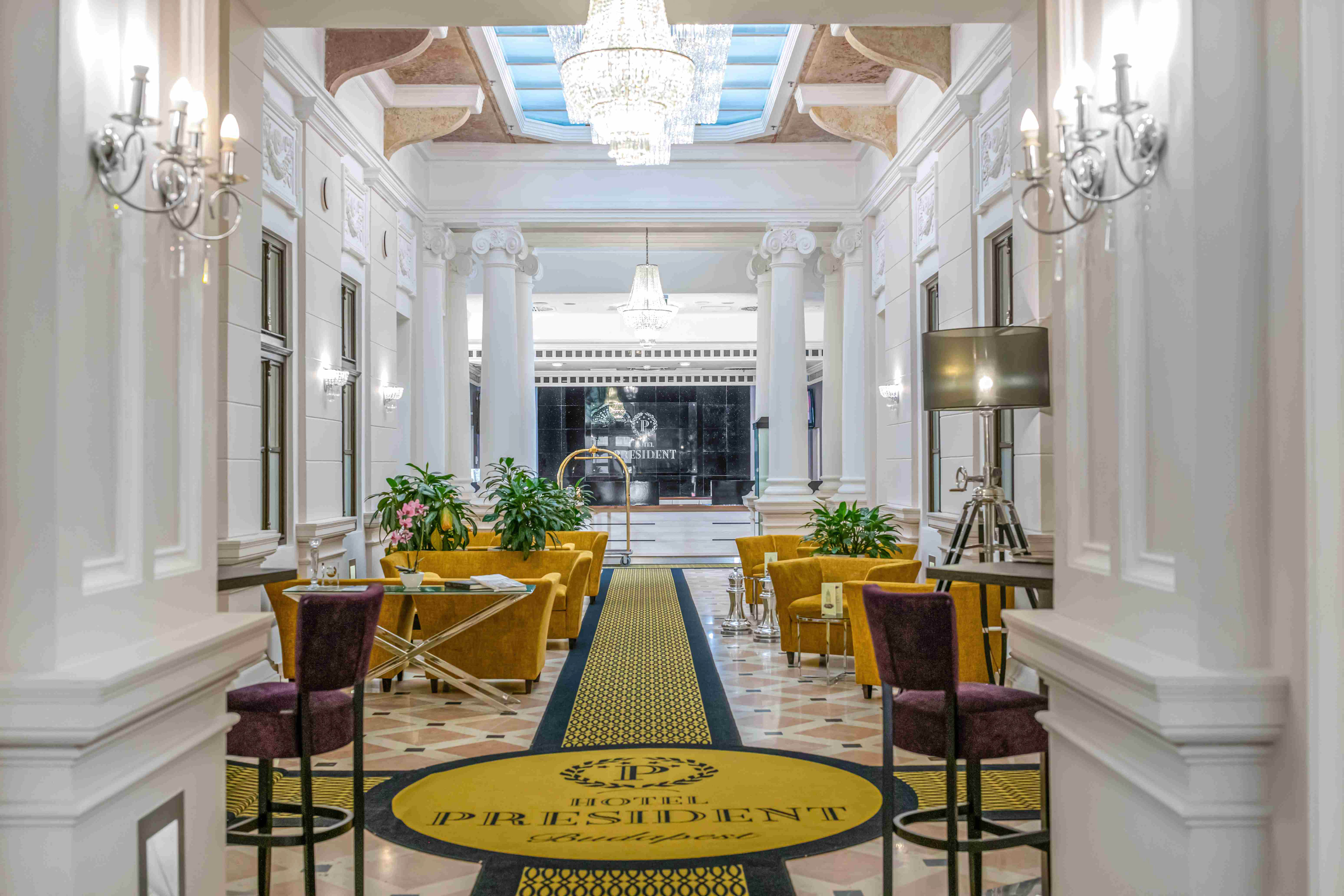 Elegant Hotel President Budapest lobby with chandeliers, columns, and gold seating.