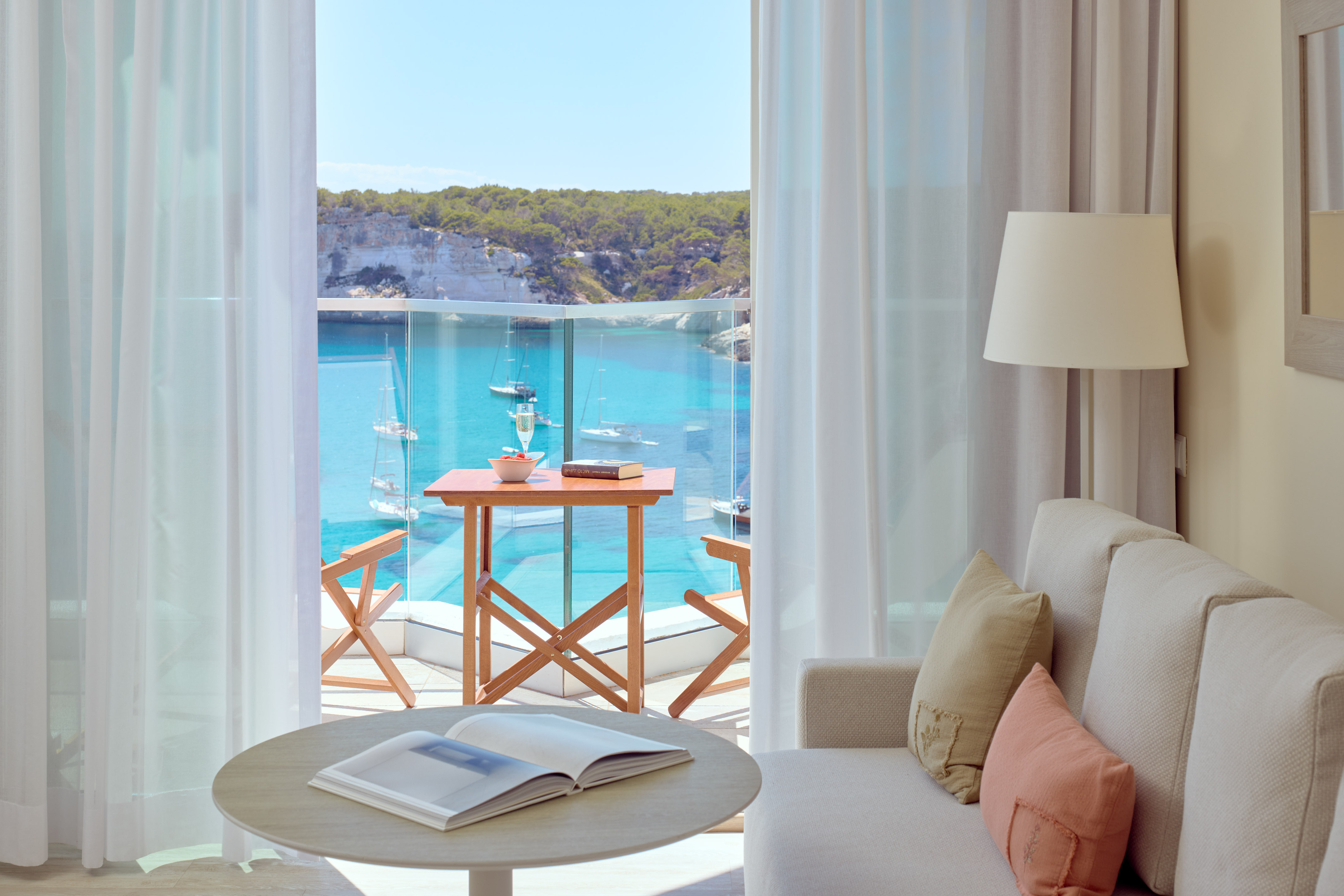 Modern hotel room with balcony overlooking turquoise sea, boats, cliffs.