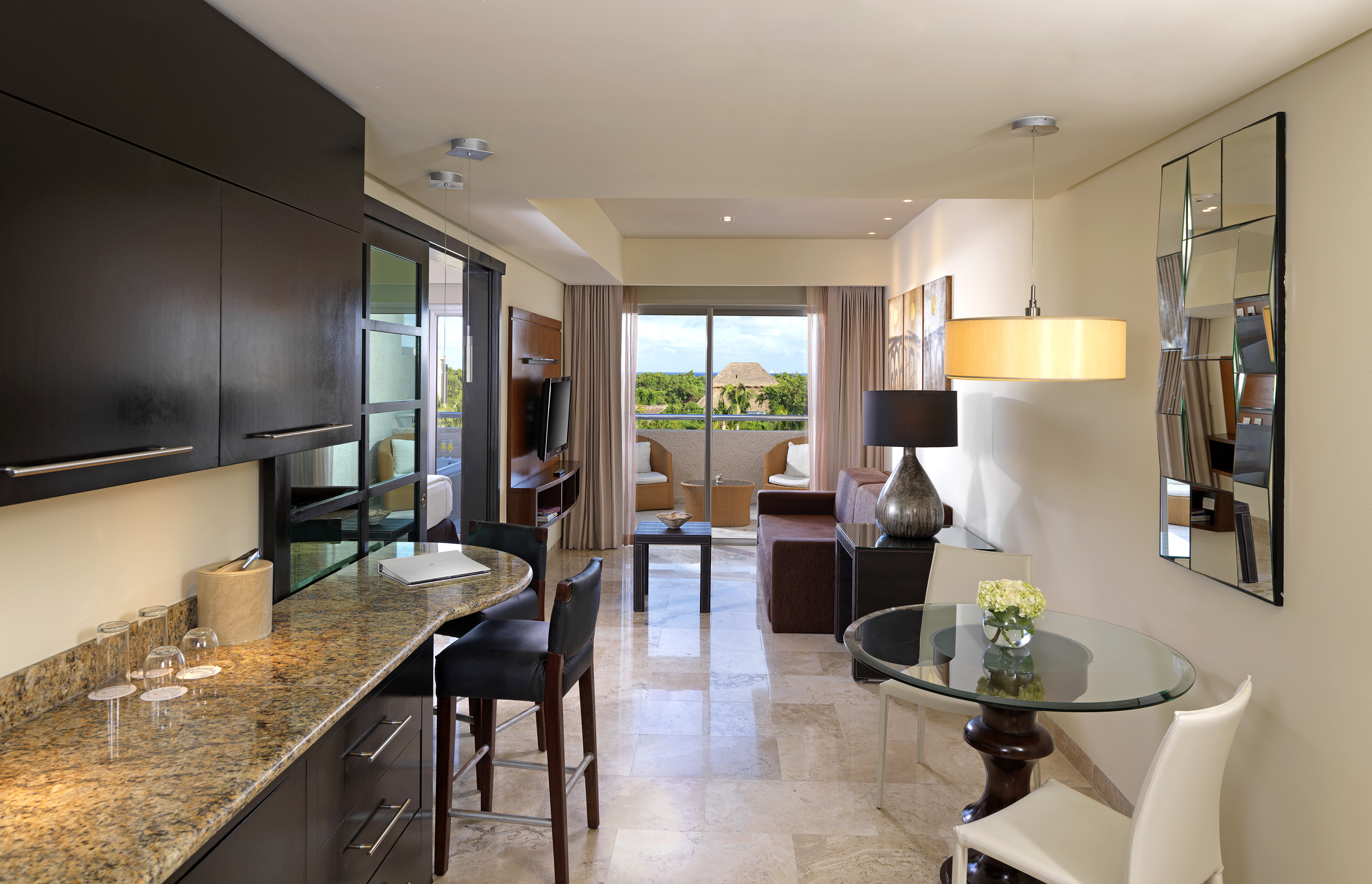 Modern hotel suite with kitchenette, living area, and tropical balcony.