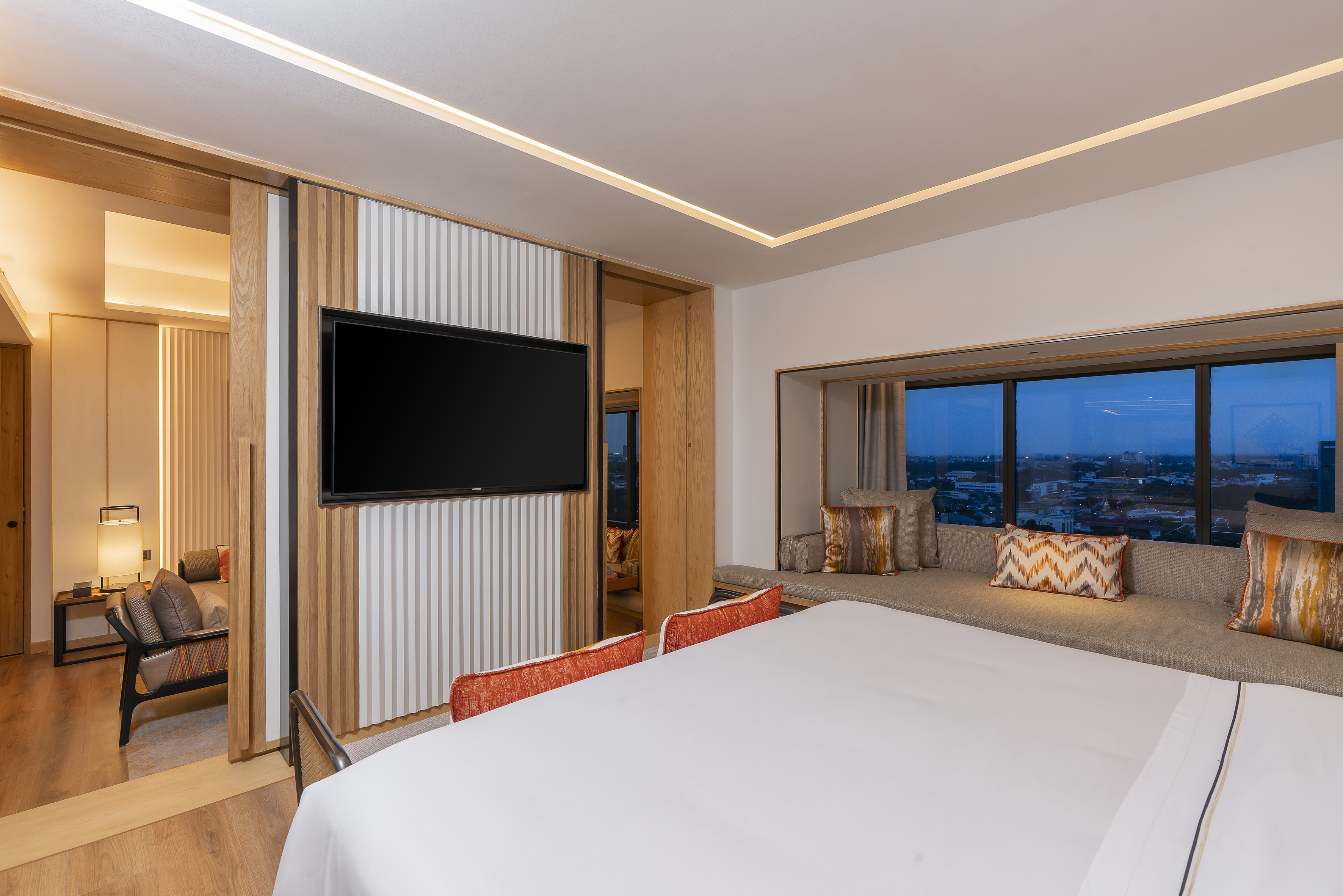 Modern hotel bedroom with white bed, mounted TV, window seat, and city view.