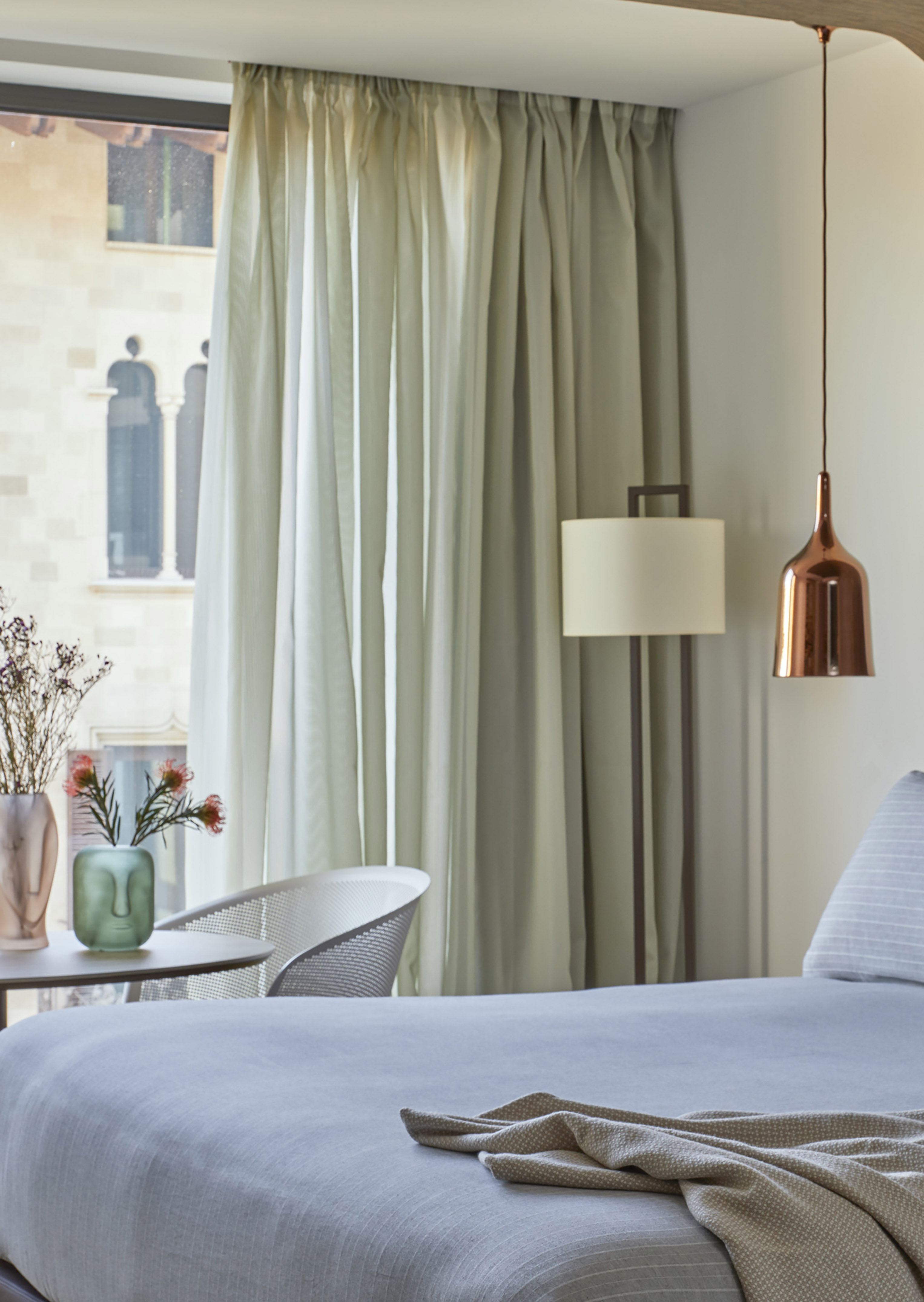 Modern hotel bedroom with light gray bed, sheer curtains, and city window view.