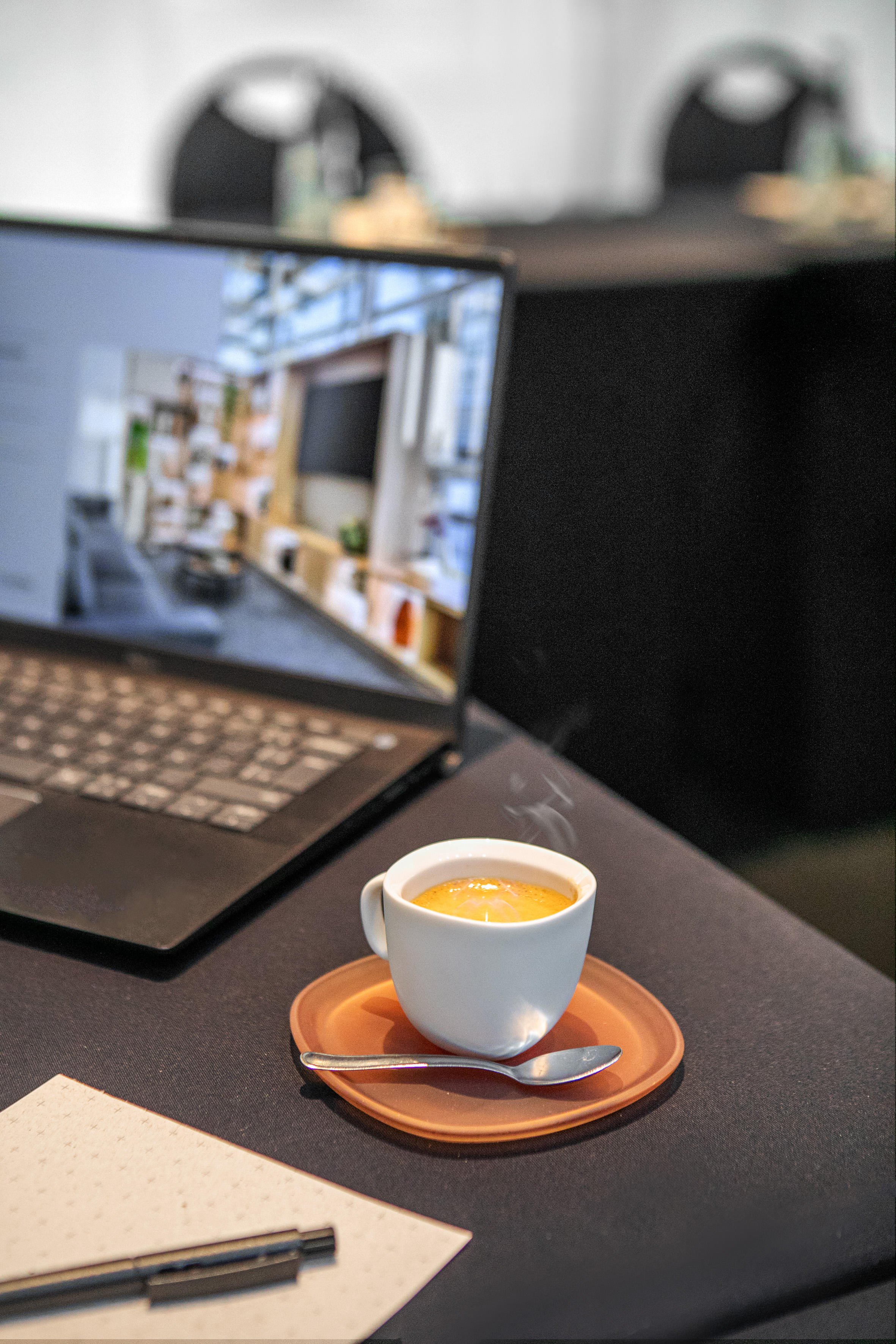 Steaming hot drink, laptop displaying interior, notebook on desk.