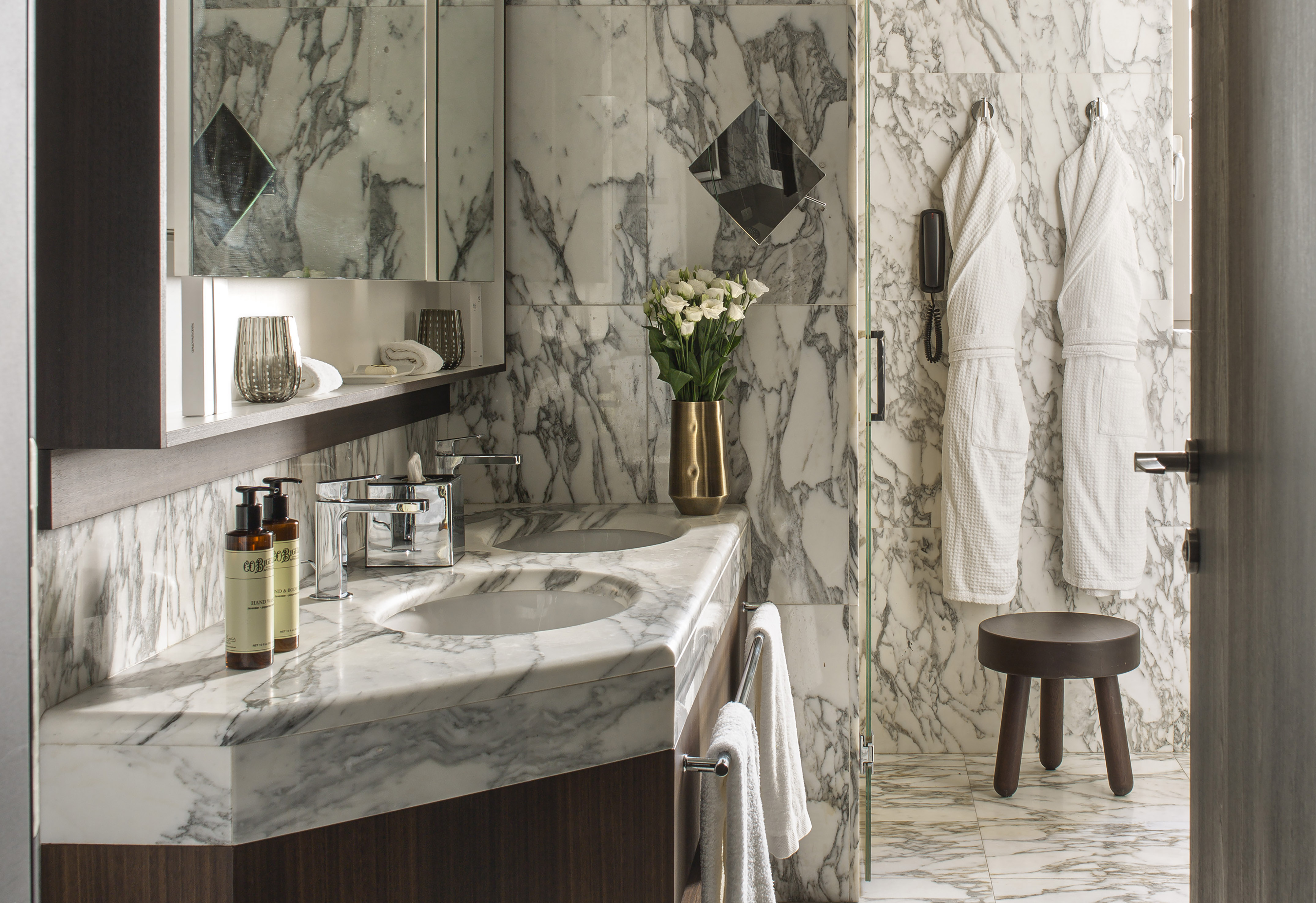 Elegant marble bathroom with double vanity, bathrobes, and white flowers.