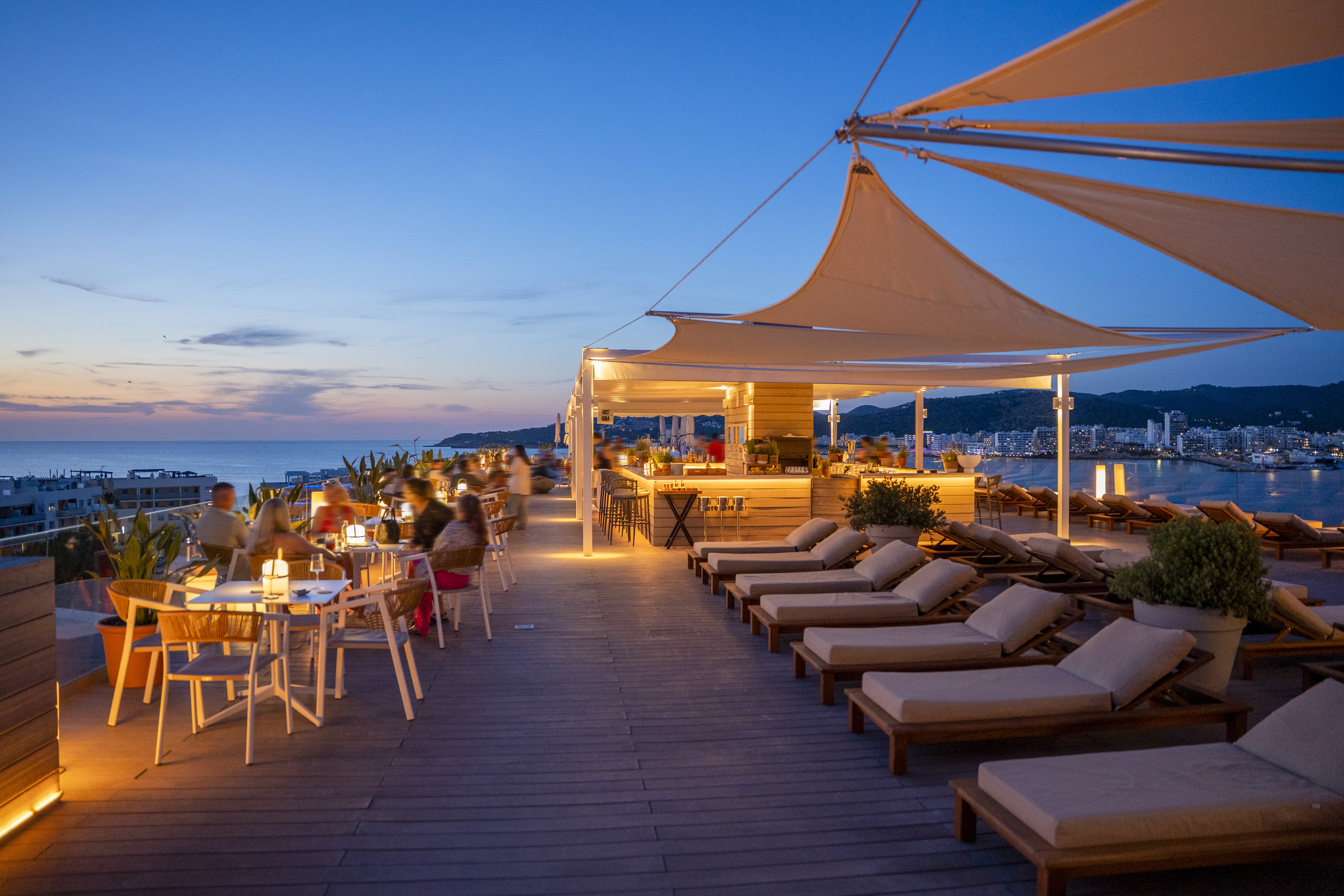 Evening rooftop bar with dining guests, lounge chairs, and ocean view.