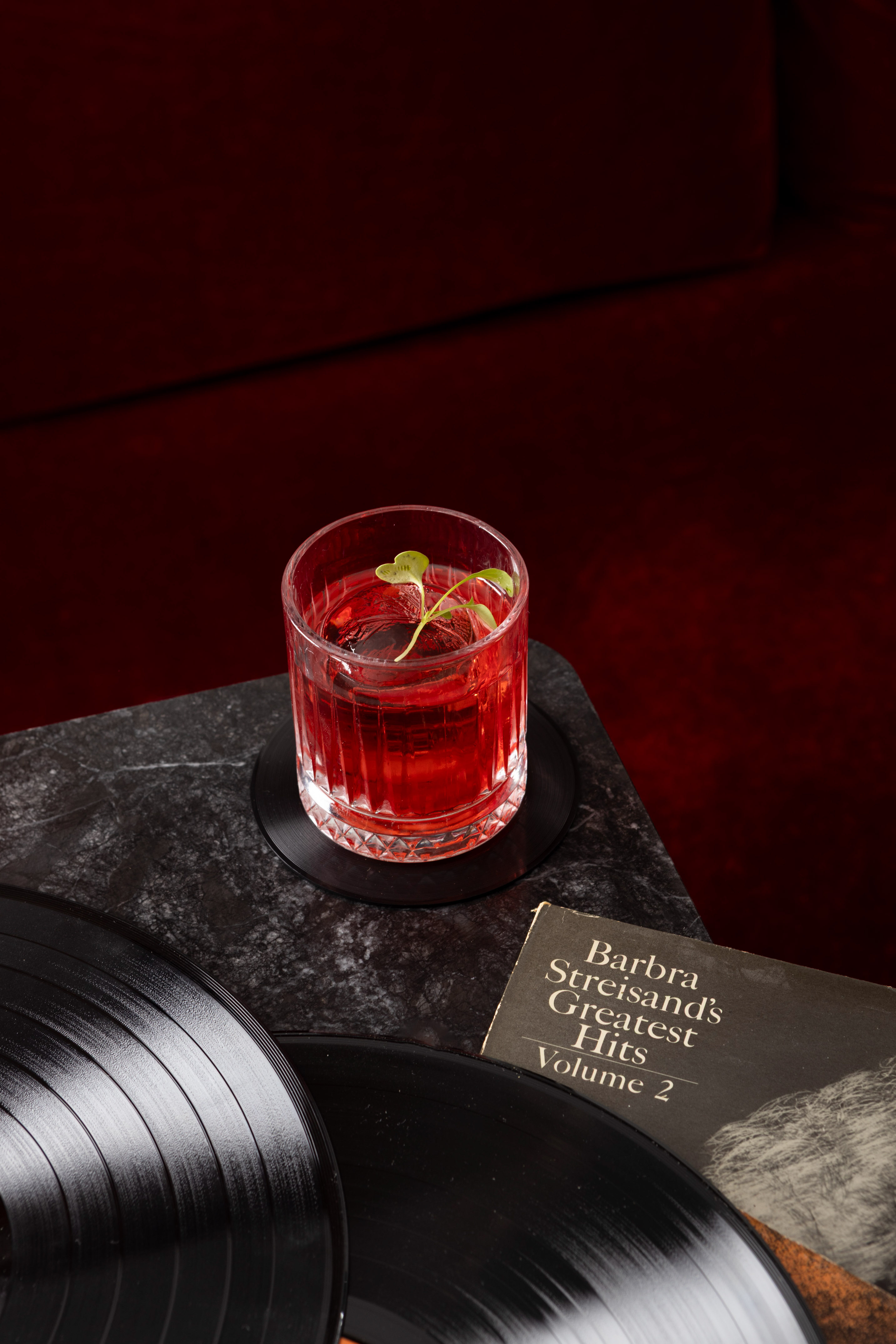 Red cocktail, vinyl records, Barbra Streisand album on marble table.