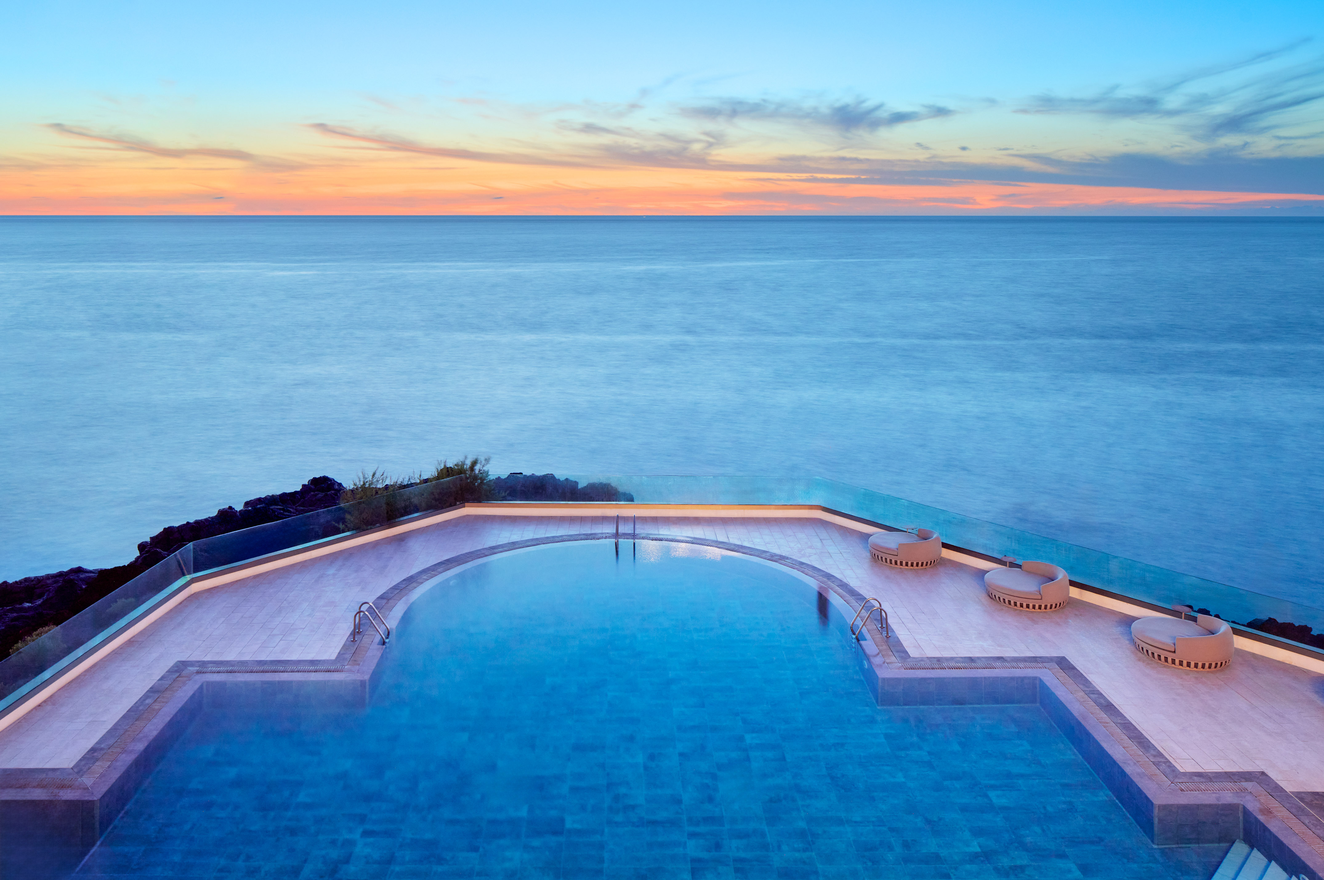 Modern infinity pool overlooking the ocean at sunset with lounge chairs.