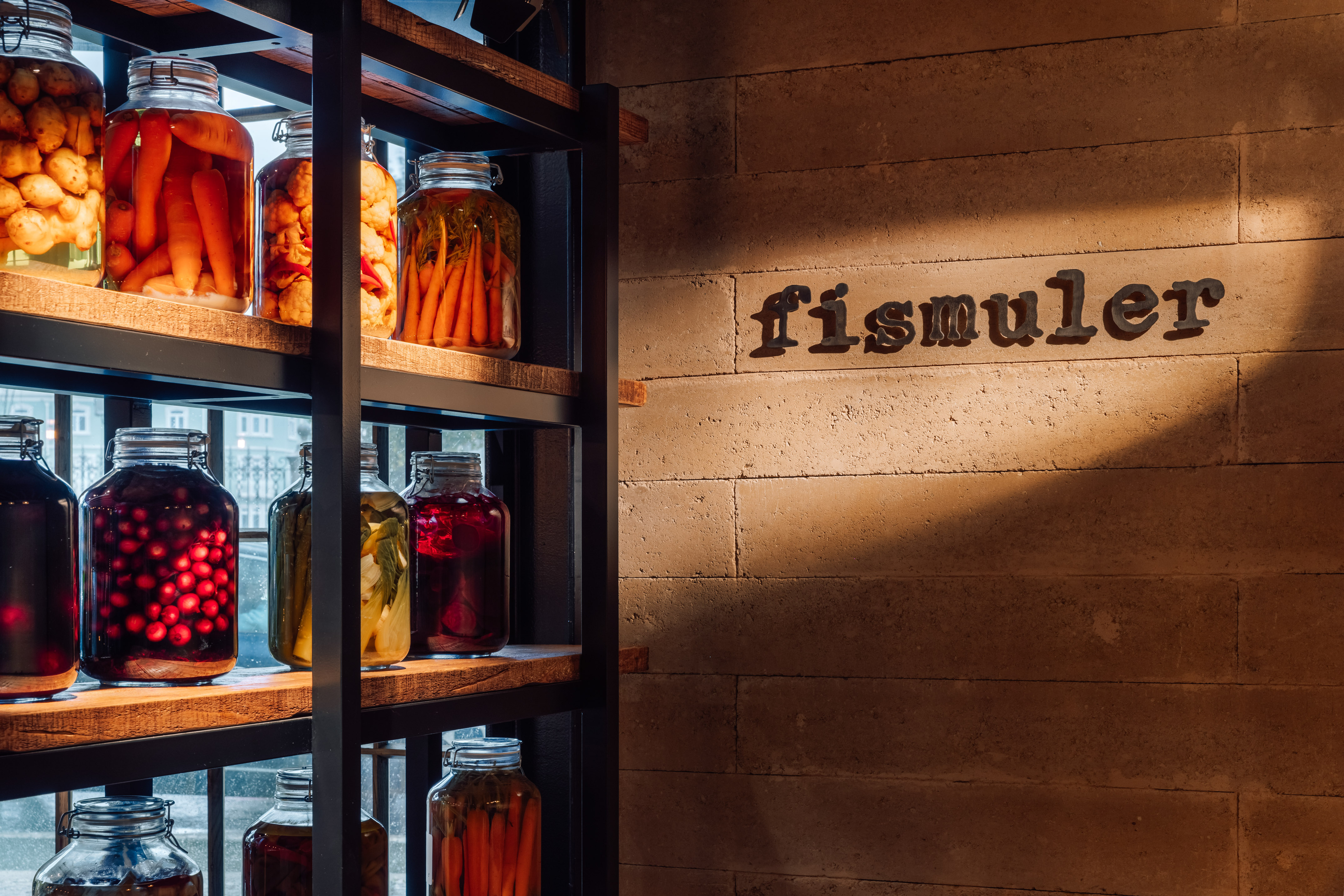 Pickled carrots, berries in jars on shelves, 'Fismuler' sign on textured wall.