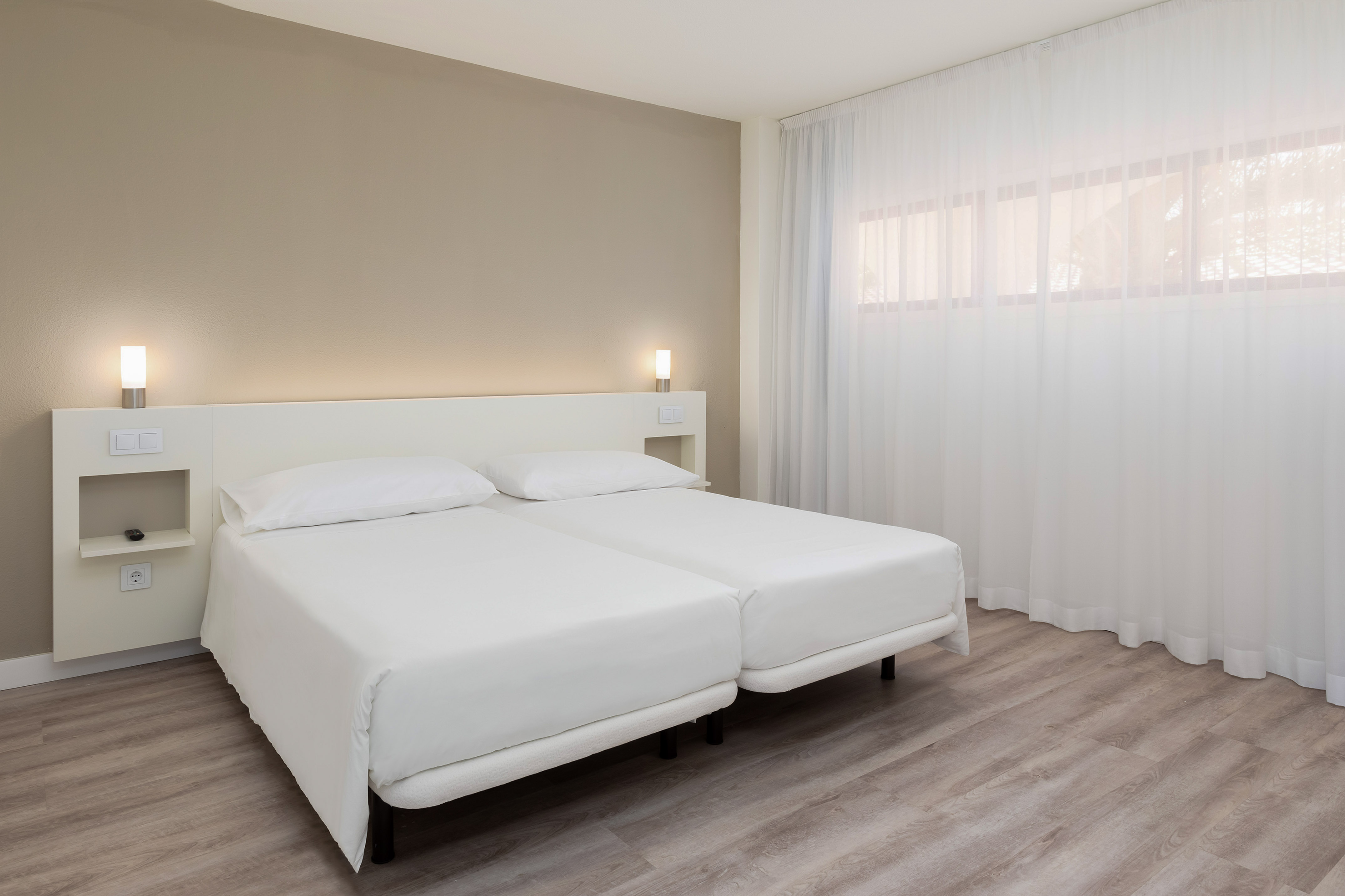 Modern hotel bedroom with twin beds, white bedding, beige wall, and wood floor.