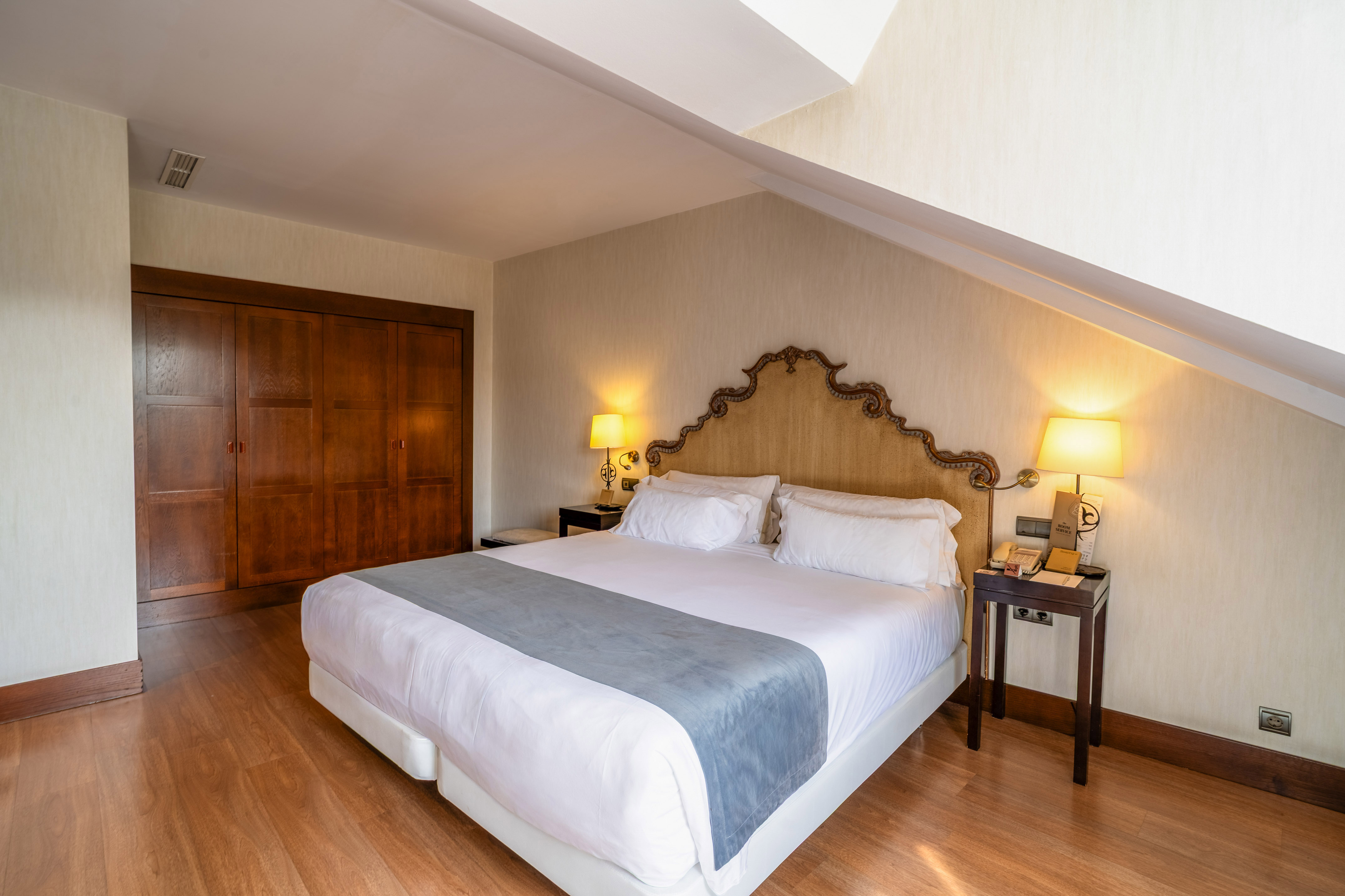 Hotel room with large bed, ornate headboard, wooden wardrobe, and sloped ceiling.