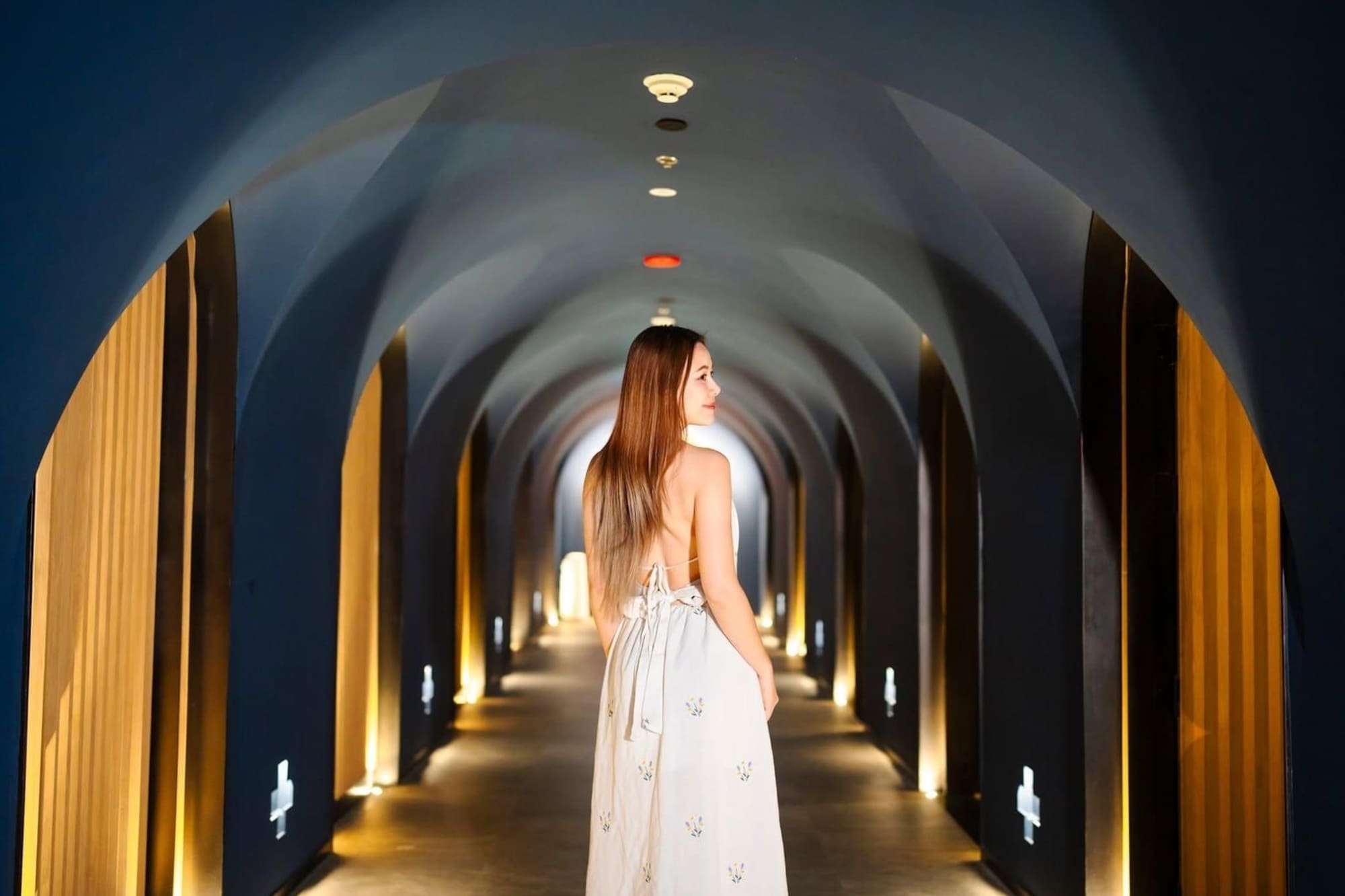 Woman in white dress looking back in modern arched hotel hallway.