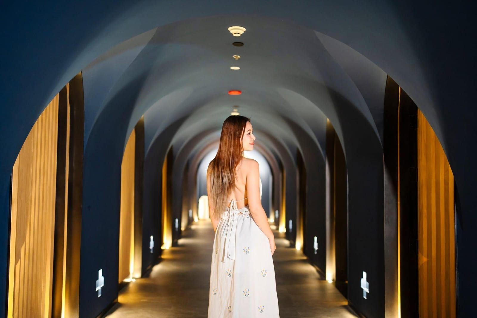 Woman in white dress looking back in modern arched hotel hallway.