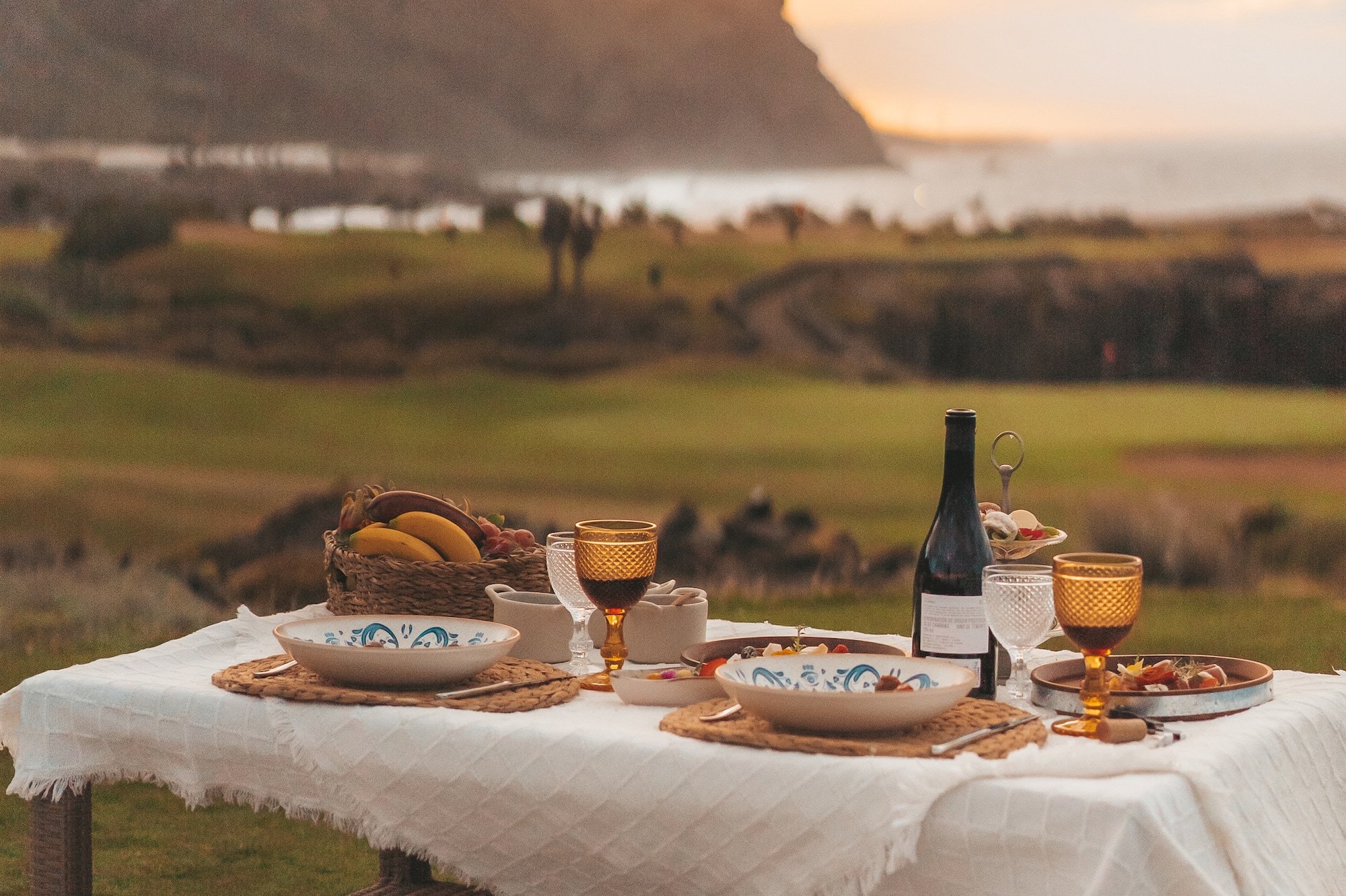 Outdoor dinner table with wine, food, and fruit, mountain and ocean sunset view.
