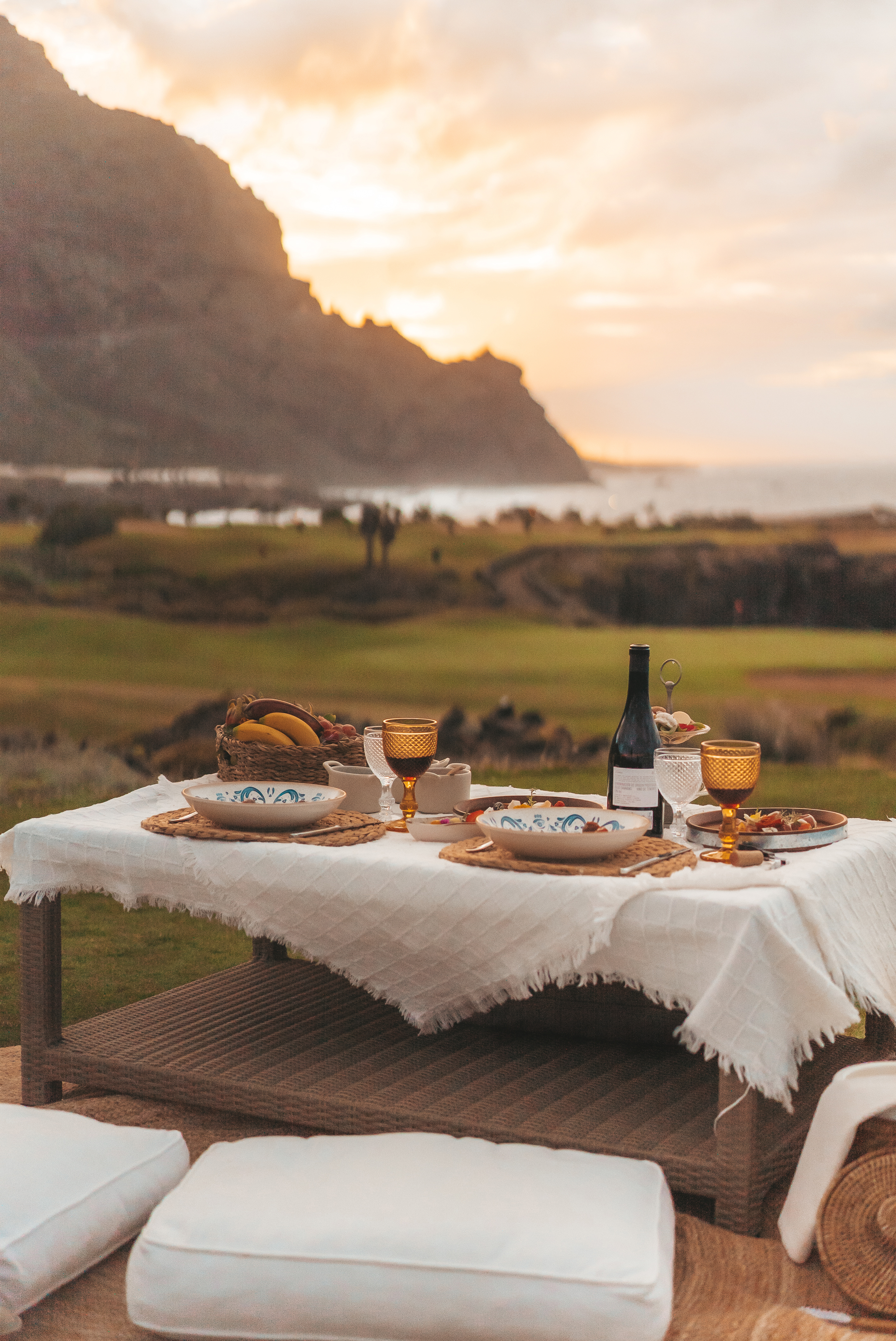 Outdoor dinner table with wine, food, and fruit, mountain and ocean sunset view.