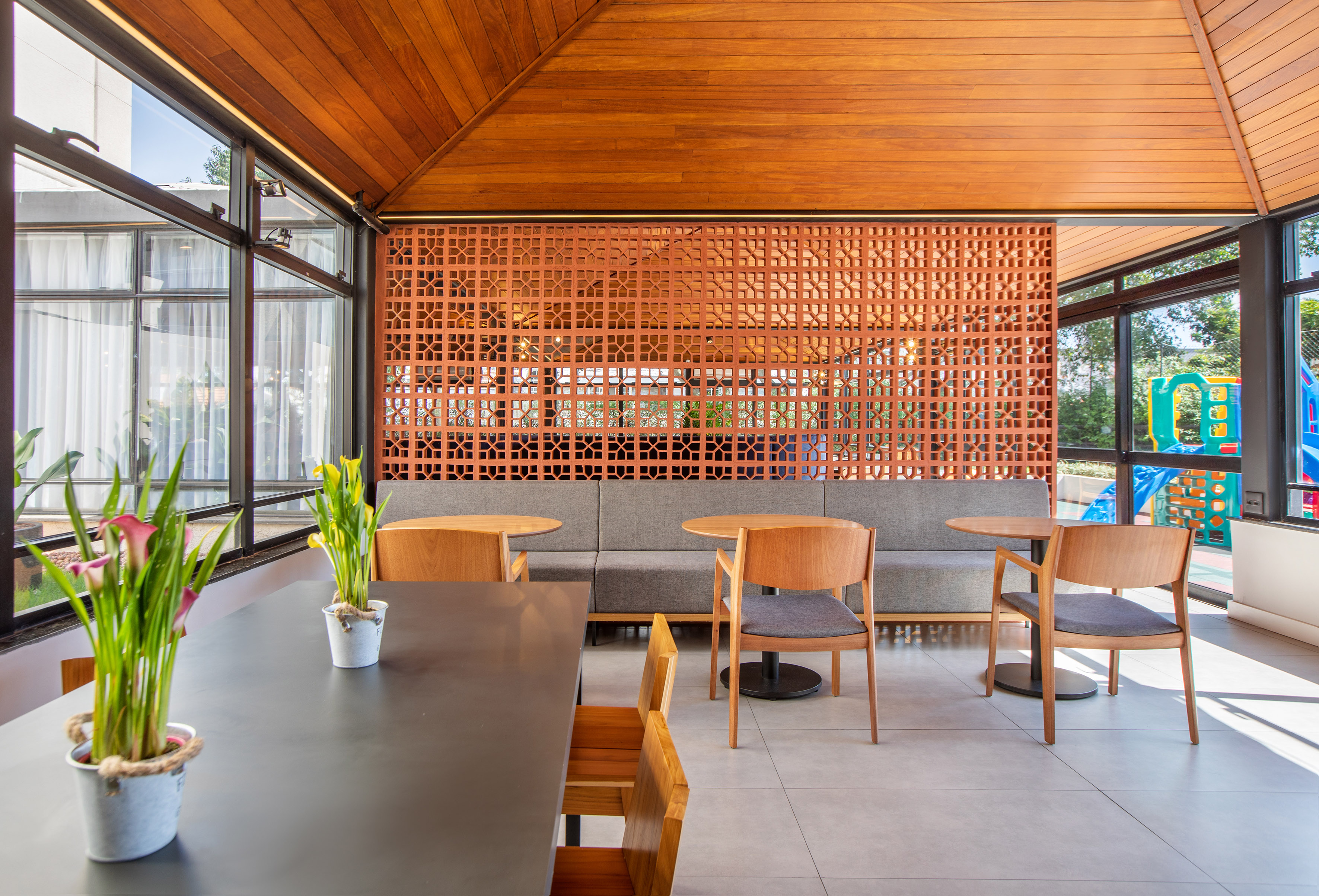 Modern lounge with wooden ceiling, terracotta screen, seating, and playground view.