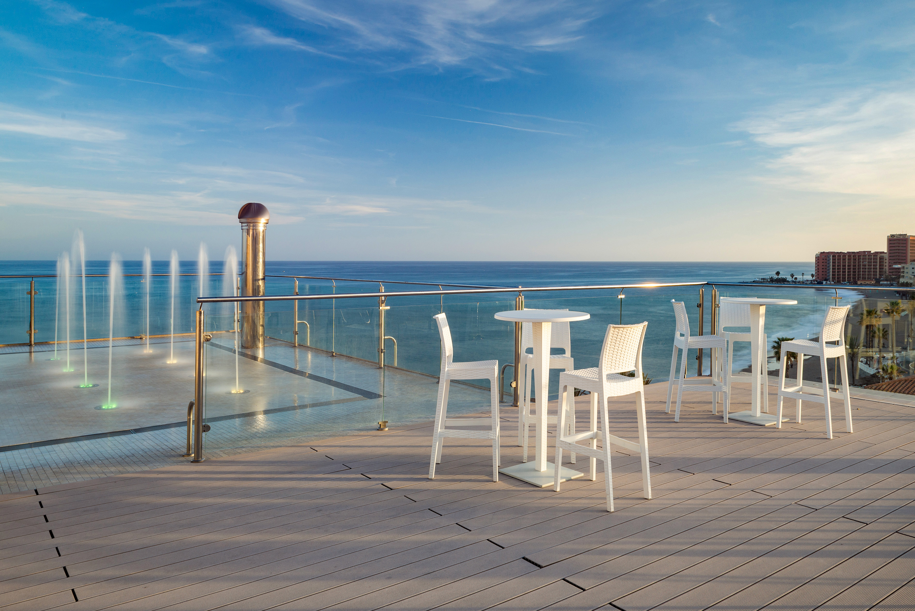 Rooftop terrace with ocean view, white bar seating, and illuminated fountains.