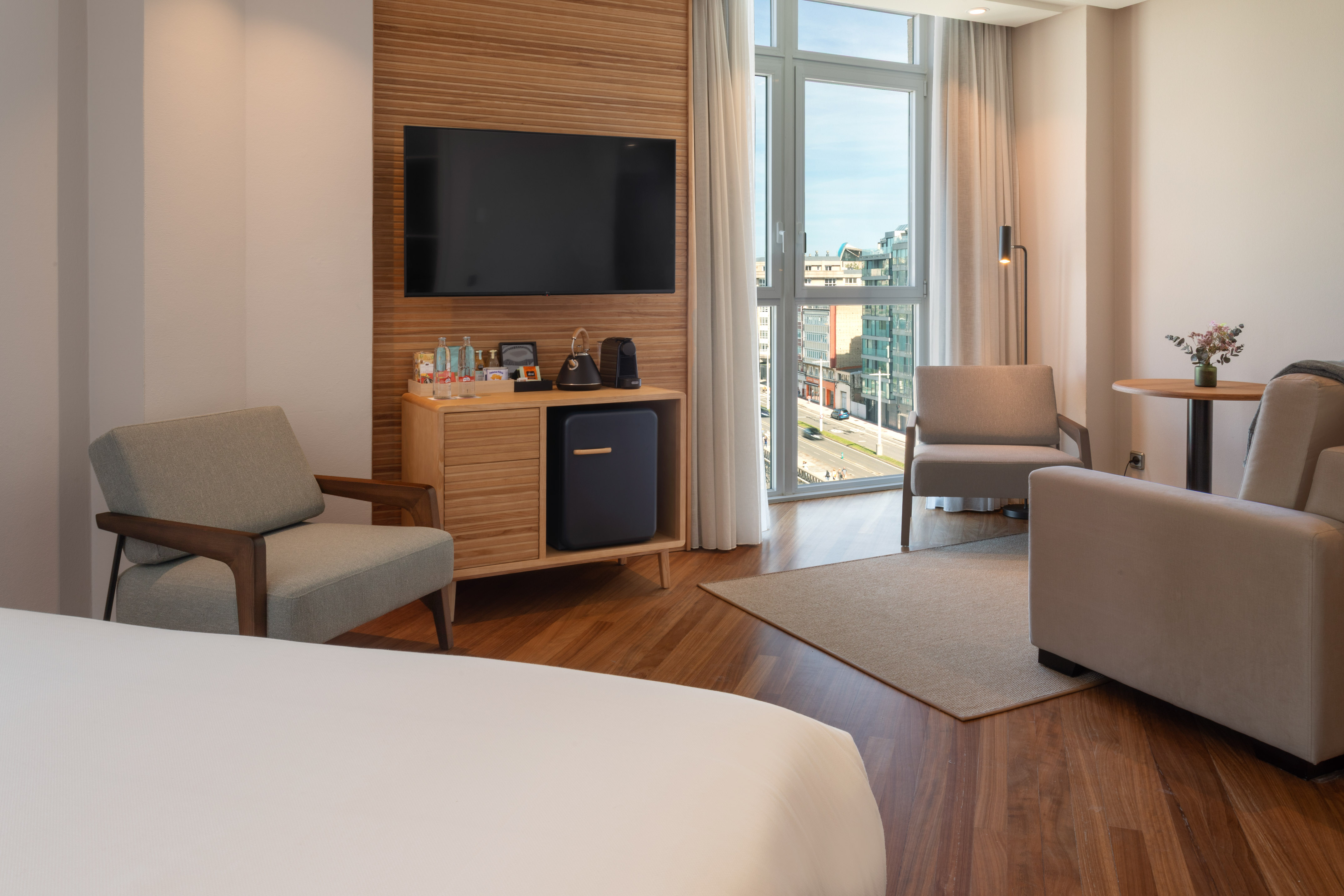 Modern hotel room with city view, TV, minibar, and two armchairs.