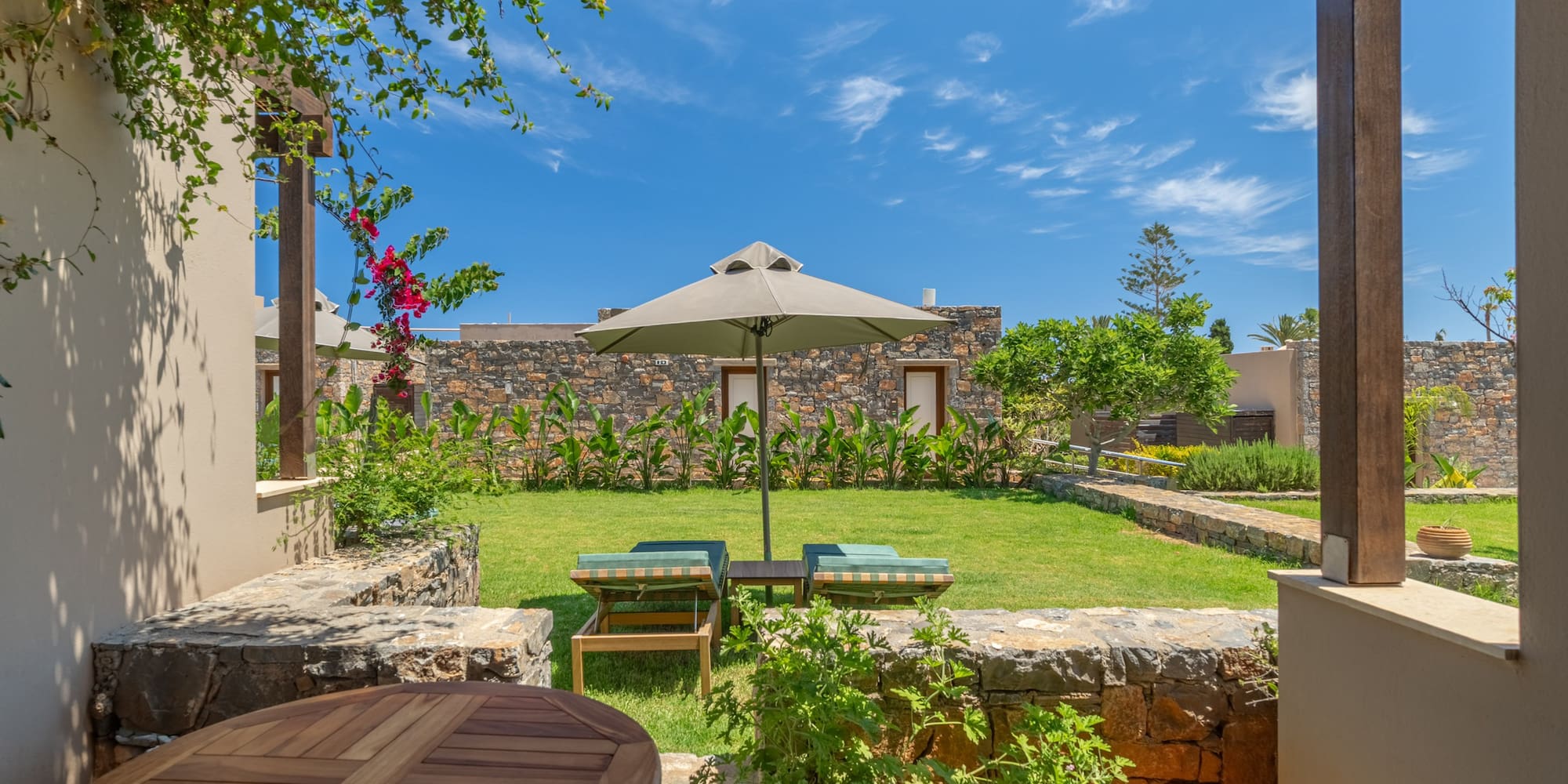 Sunny resort garden patio with loungers, parasol, and stone buildings.