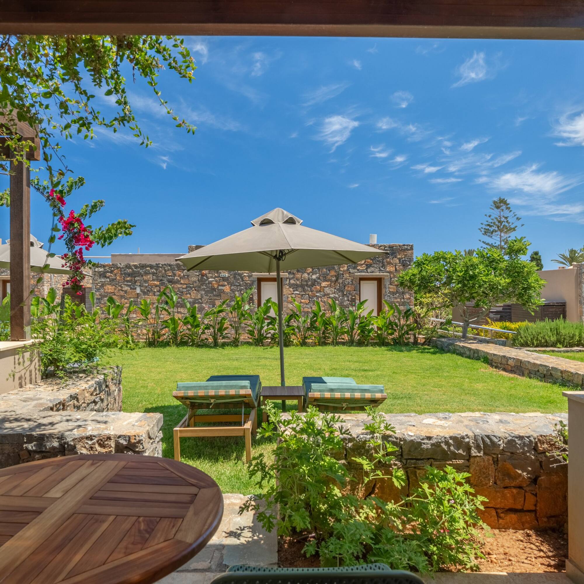 Sunny resort garden patio with loungers, parasol, and stone buildings.
