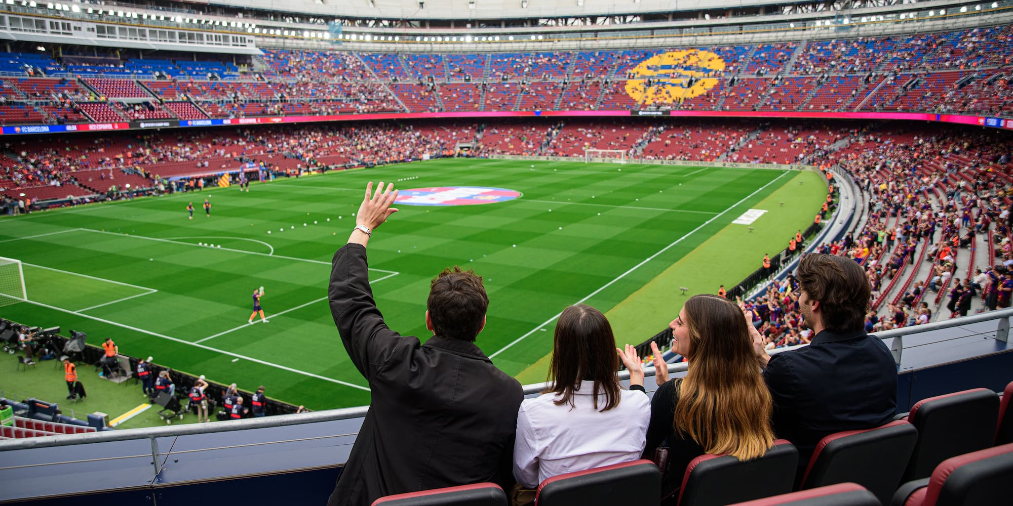 Four fans cheering in a stadium, watching a football match from seats.