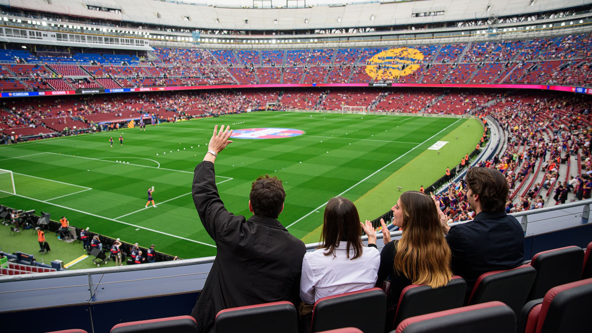 Group of friends cheering from stadium stands overlooking a vast soccer pitch.