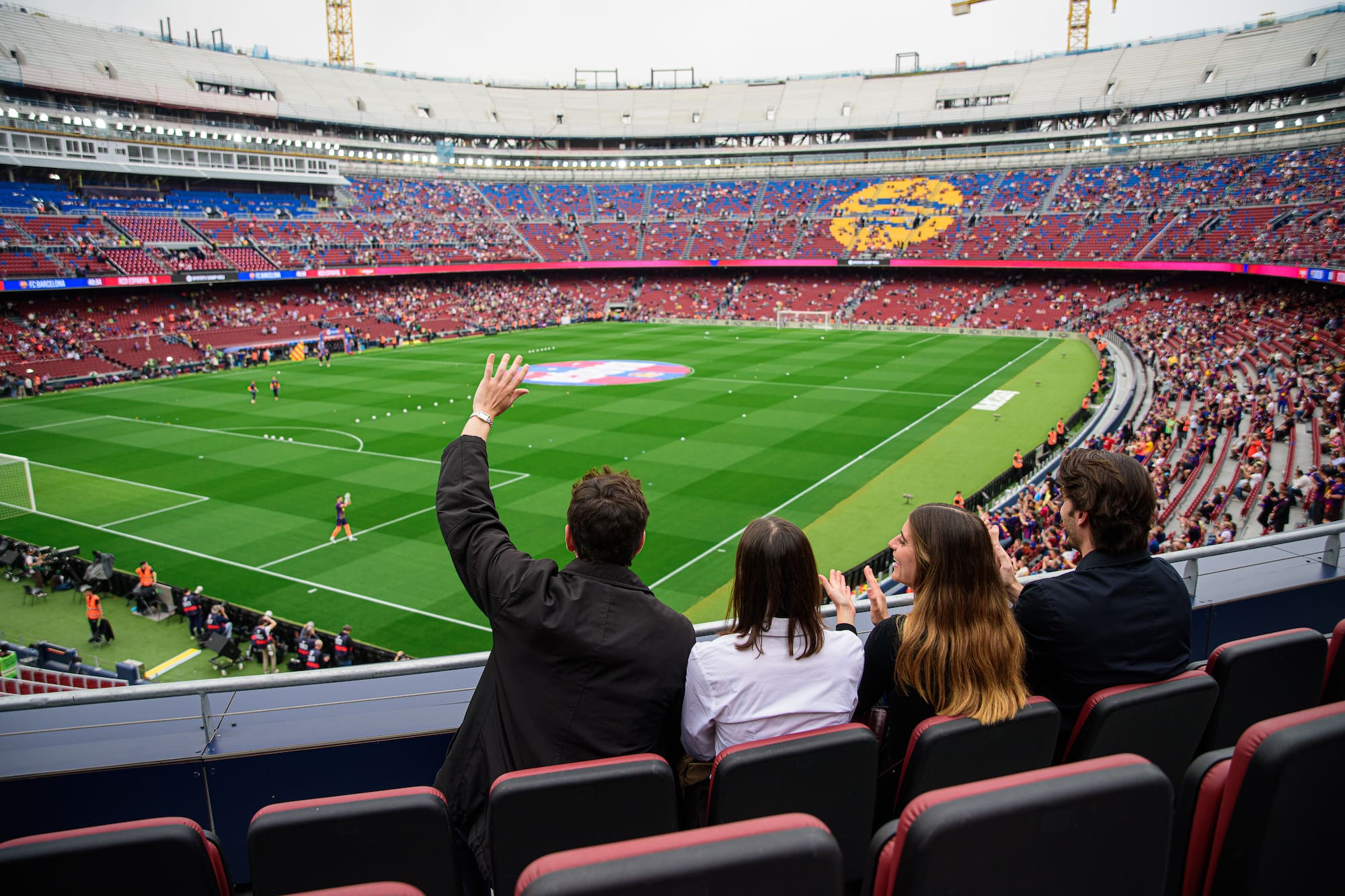 Four fans cheering in a stadium, watching a football match from seats.