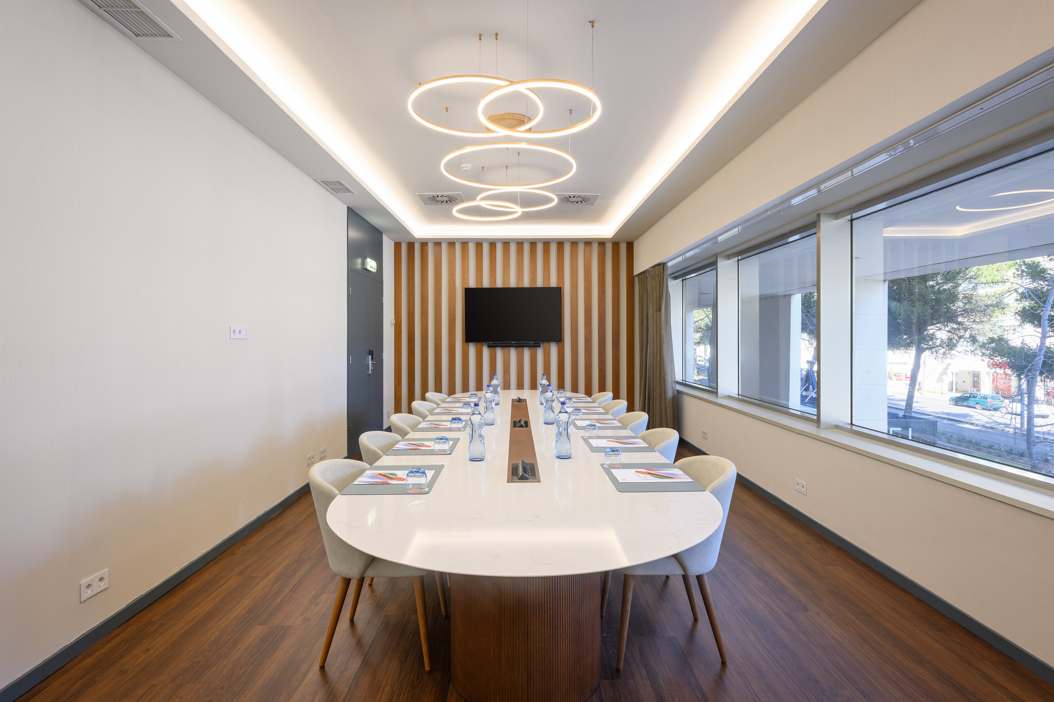 Modern conference room with oval table, circular lighting, large TV, and windows.
