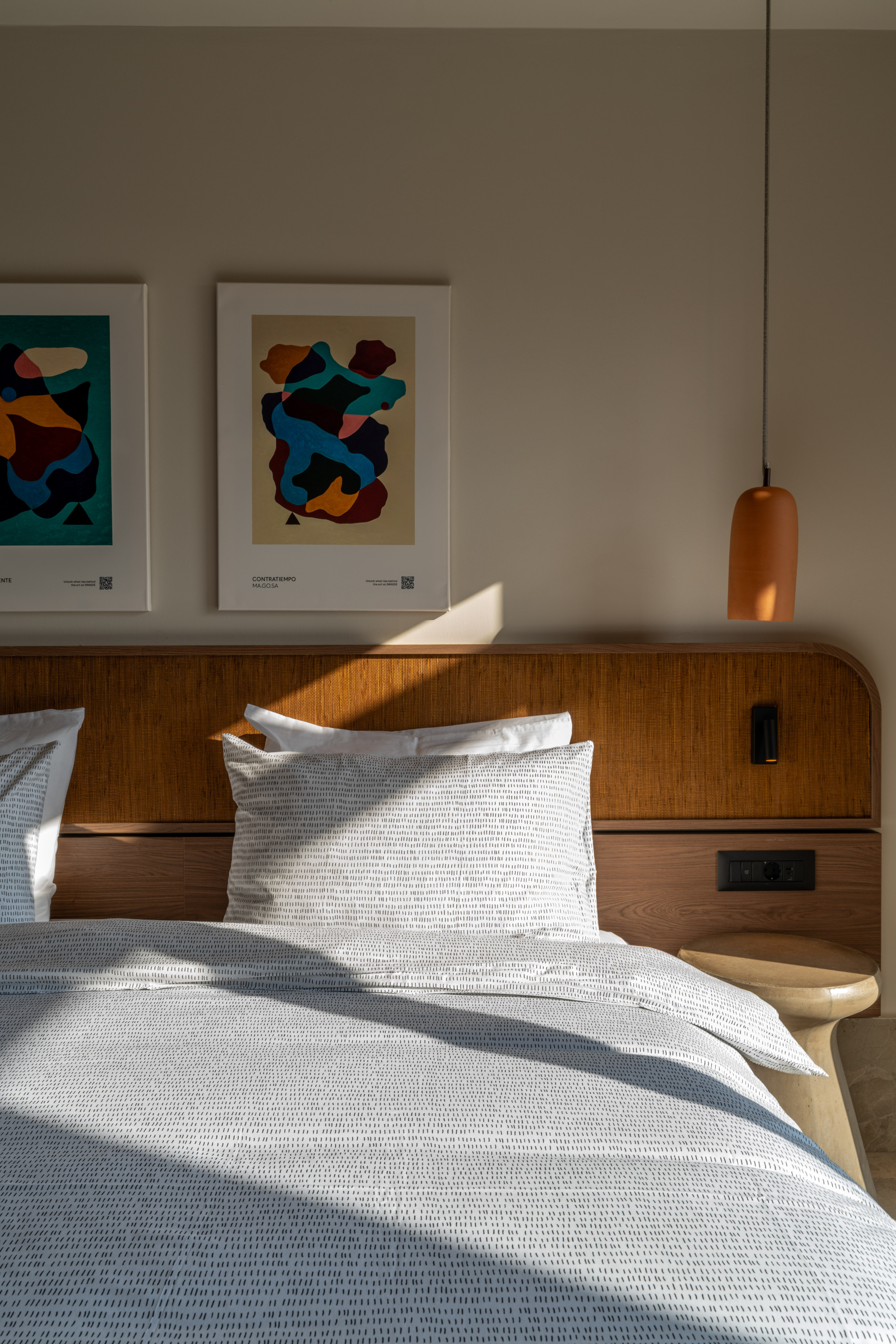 Sunlit modern bedroom with white patterned bedding, wood headboard, and abstract art.