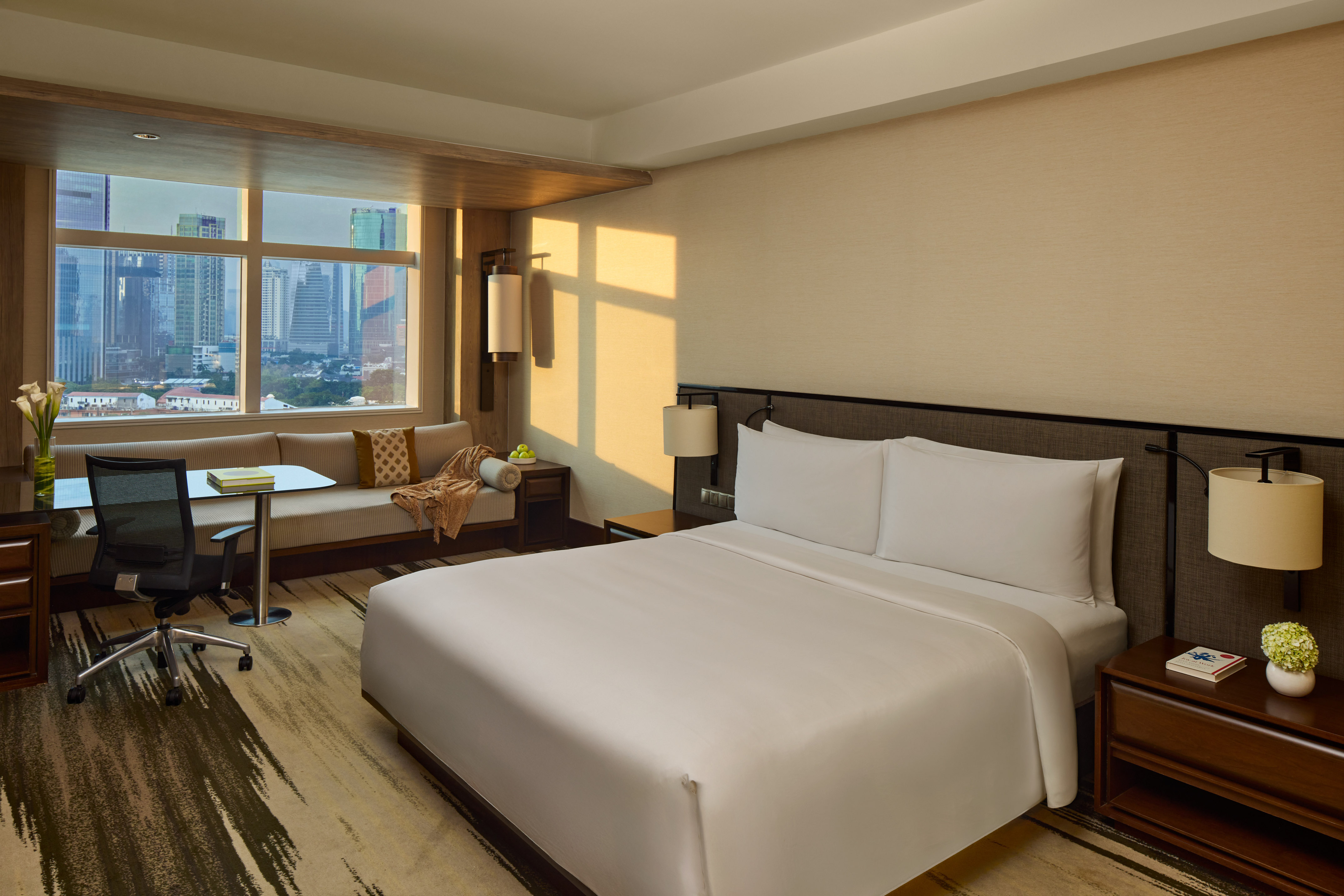 Bright hotel room with city skyline view, large bed, desk, and sofa.
