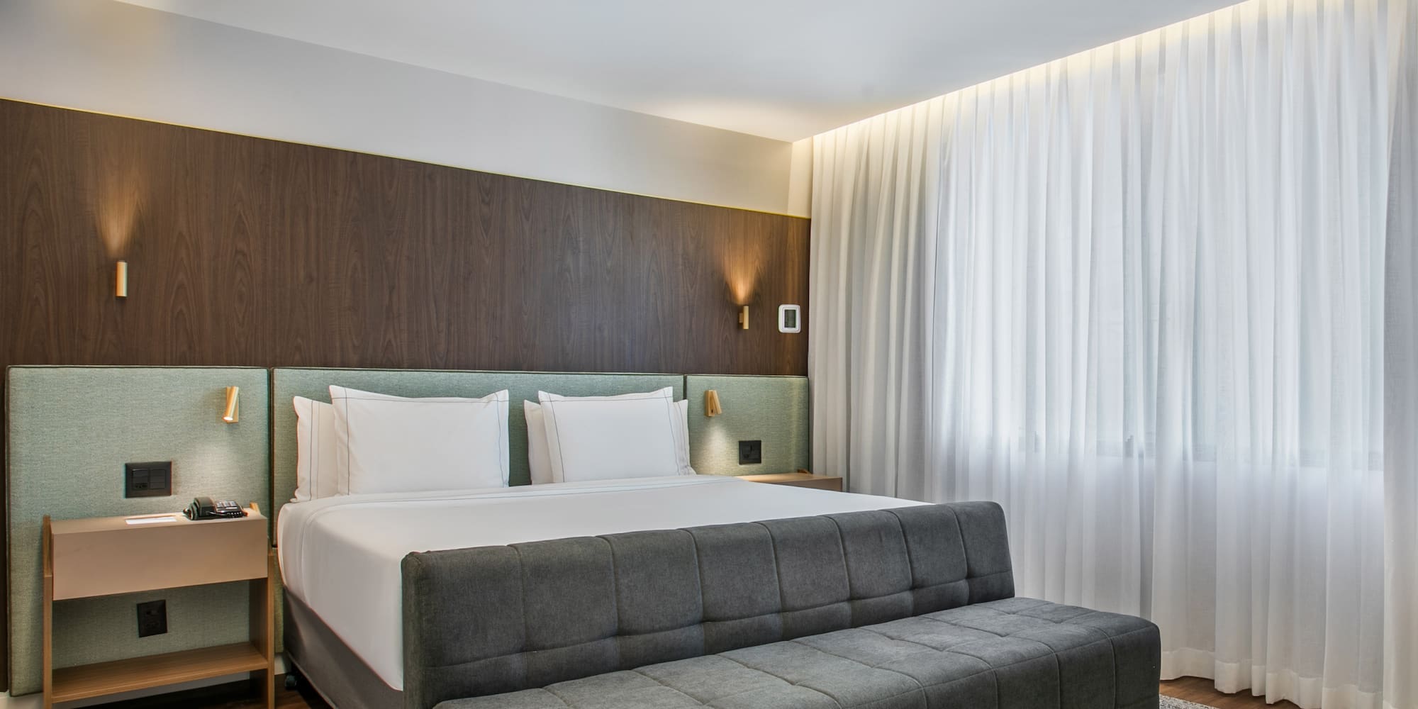 Modern hotel bedroom with king bed, wood paneling, and sheer white curtains.