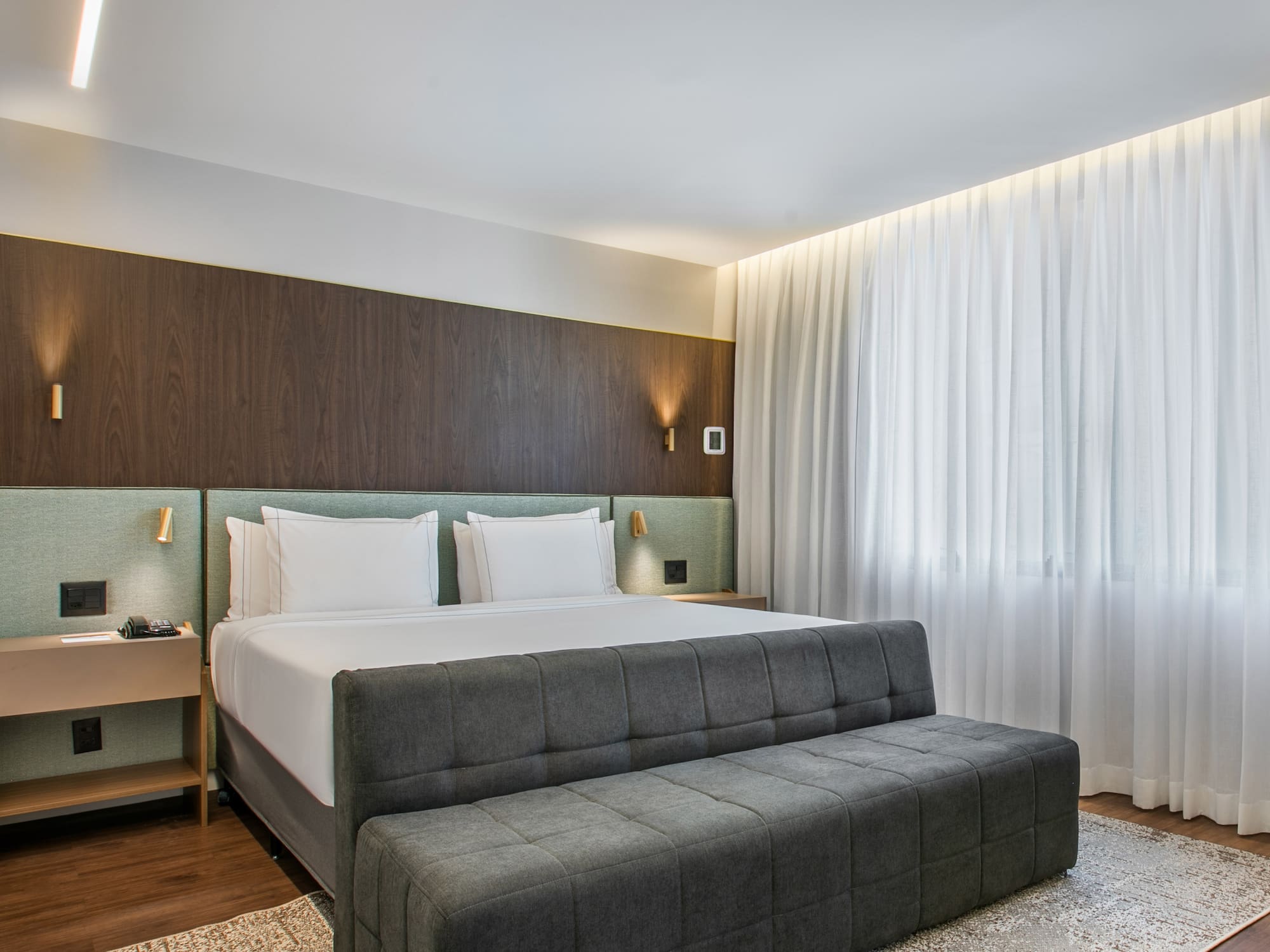 Modern hotel bedroom with king bed, wood paneling, and sheer white curtains.