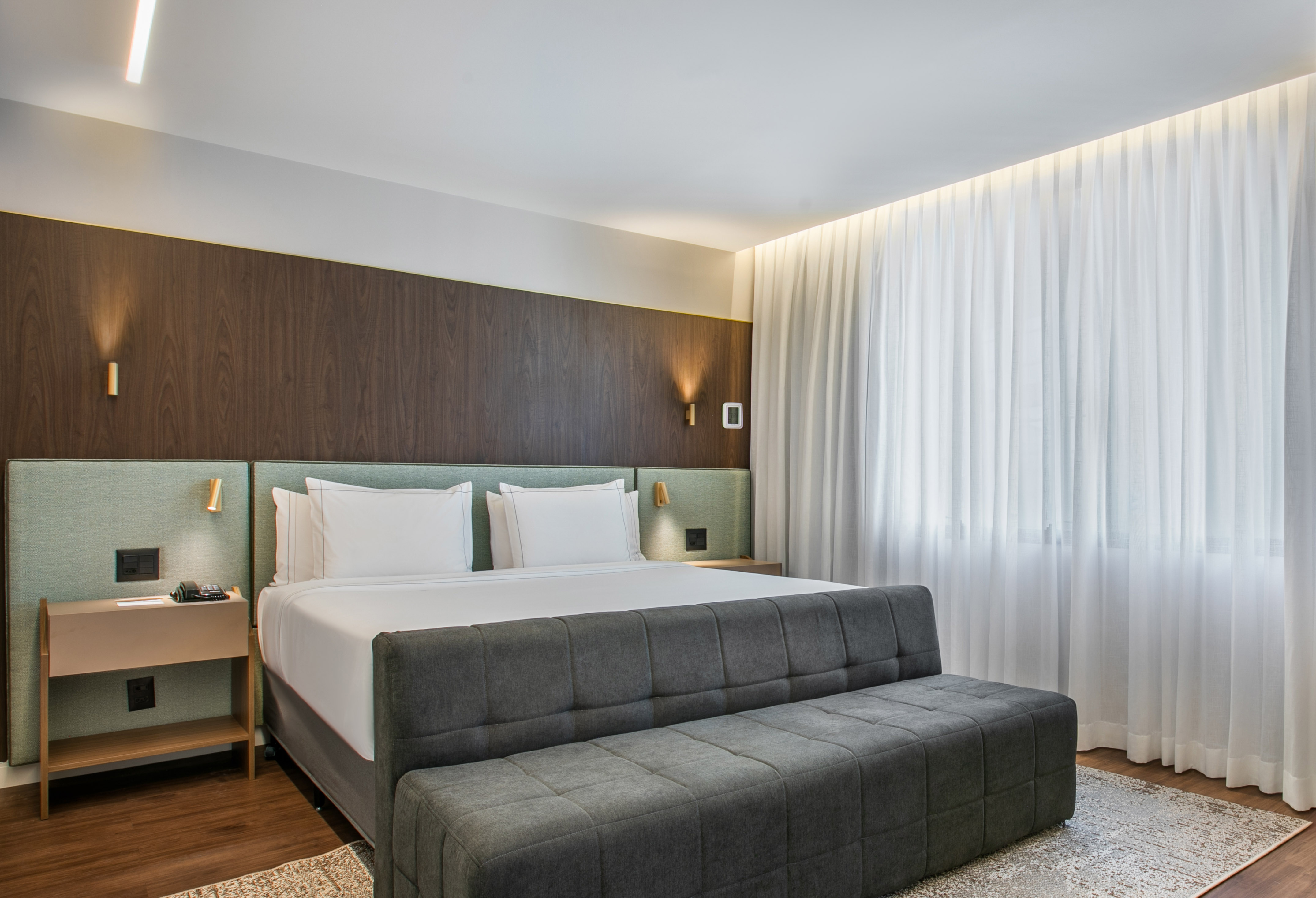 Modern hotel bedroom with king bed, wood paneling, and sheer white curtains.