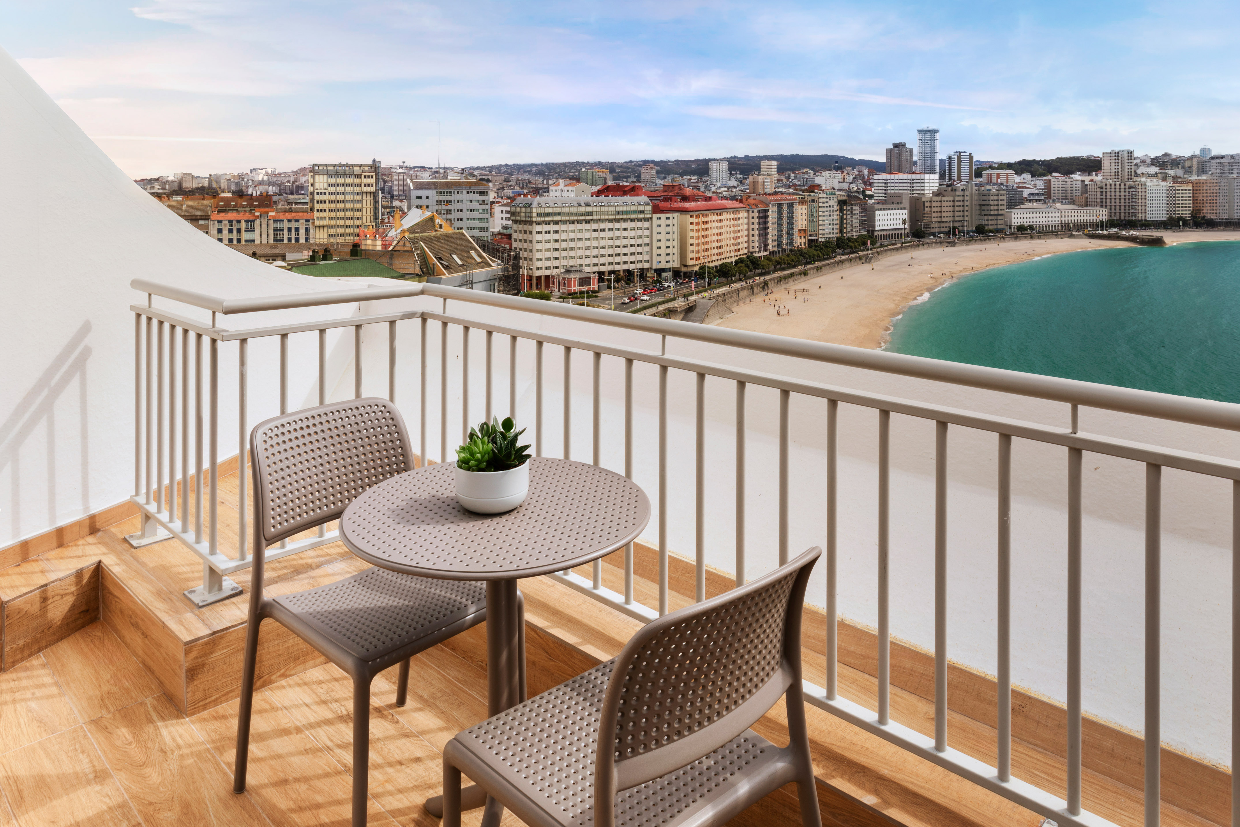 Balcón hotel con mesa y sillas, vista panorámica a playa y ciudad de A Coruña.