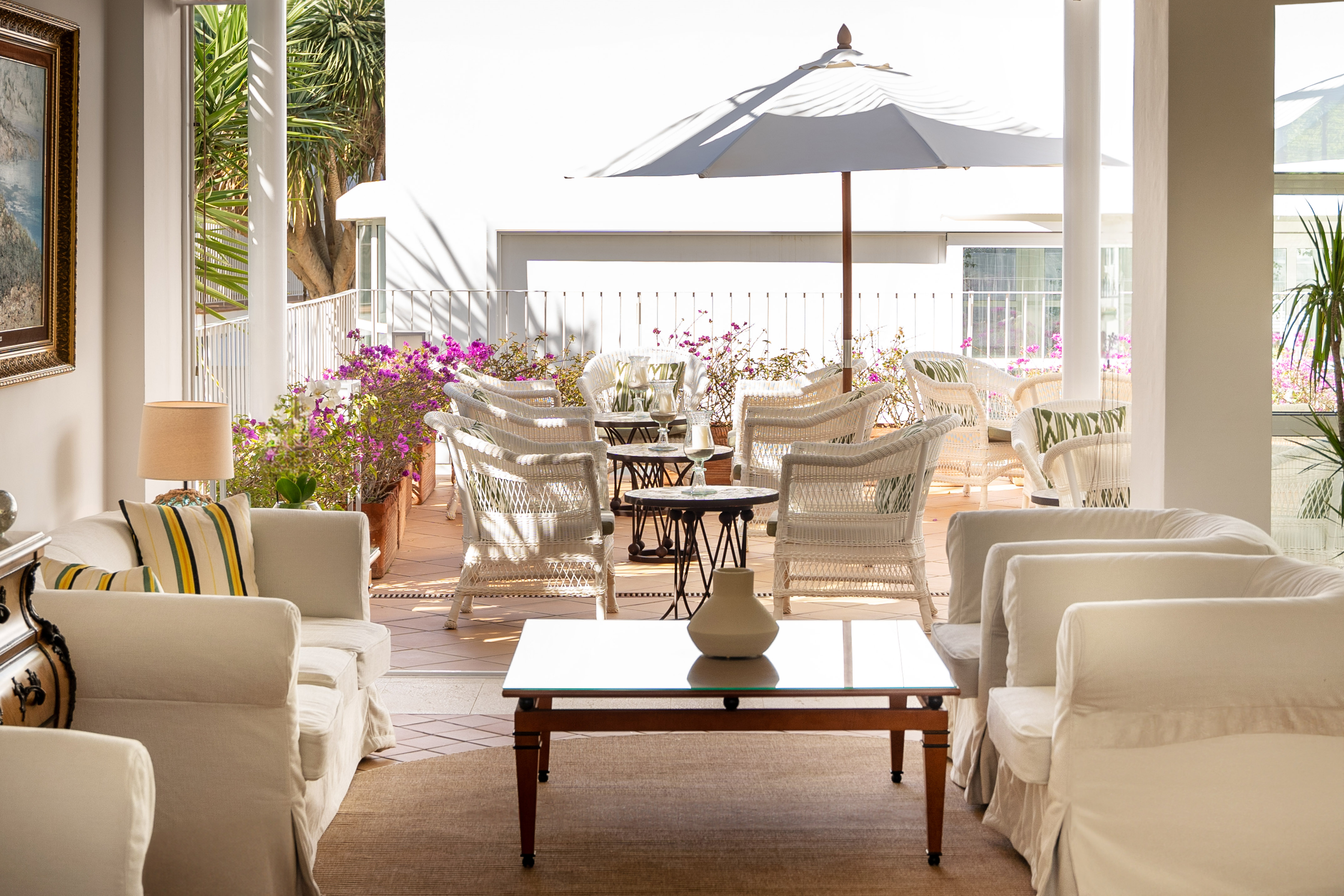 Salón y terraza de hotel con sillones de mimbre, sombrilla y plantas.