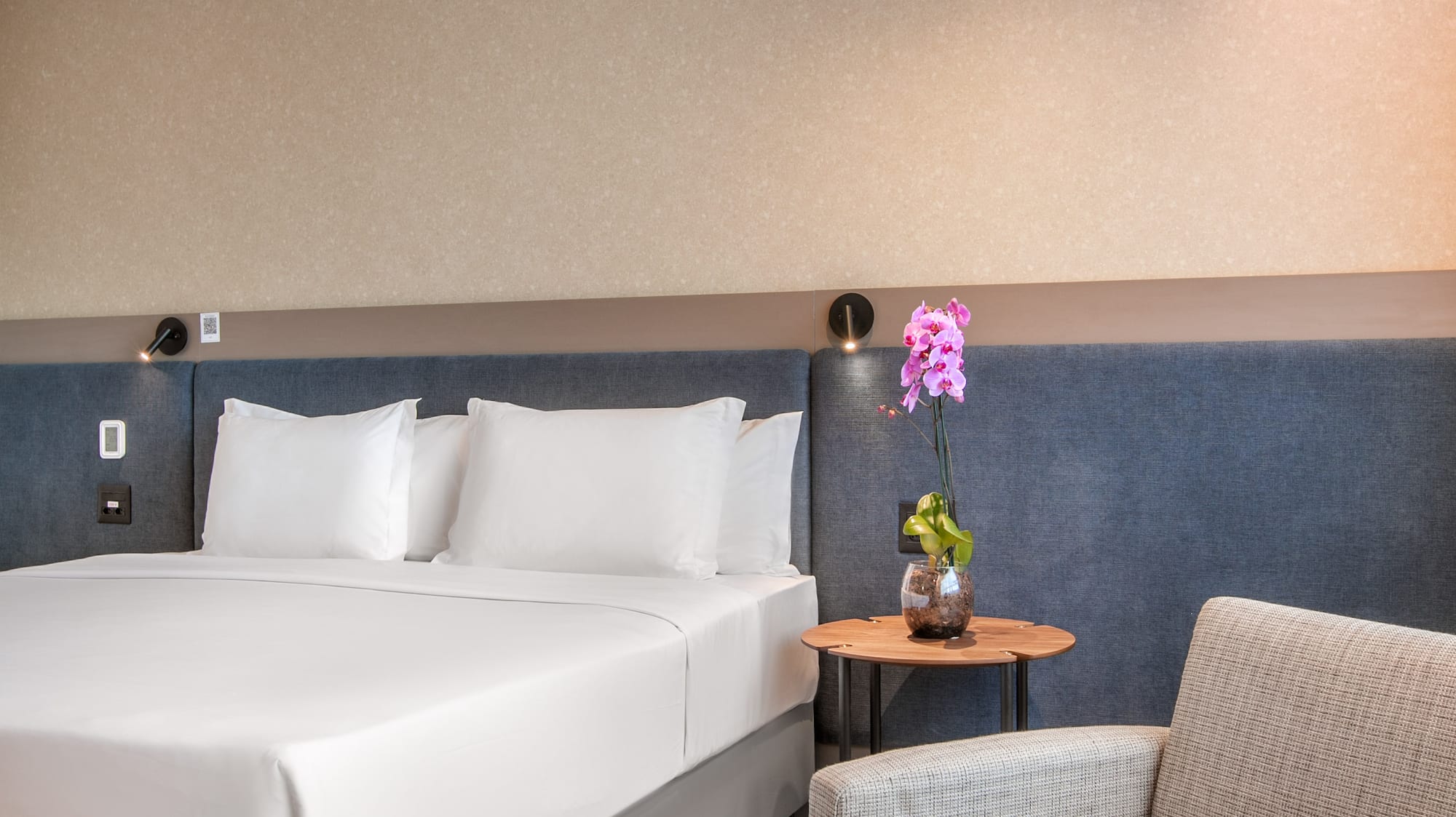 Contemporary hotel room with white bed, cozy armchair, and decorative orchid.