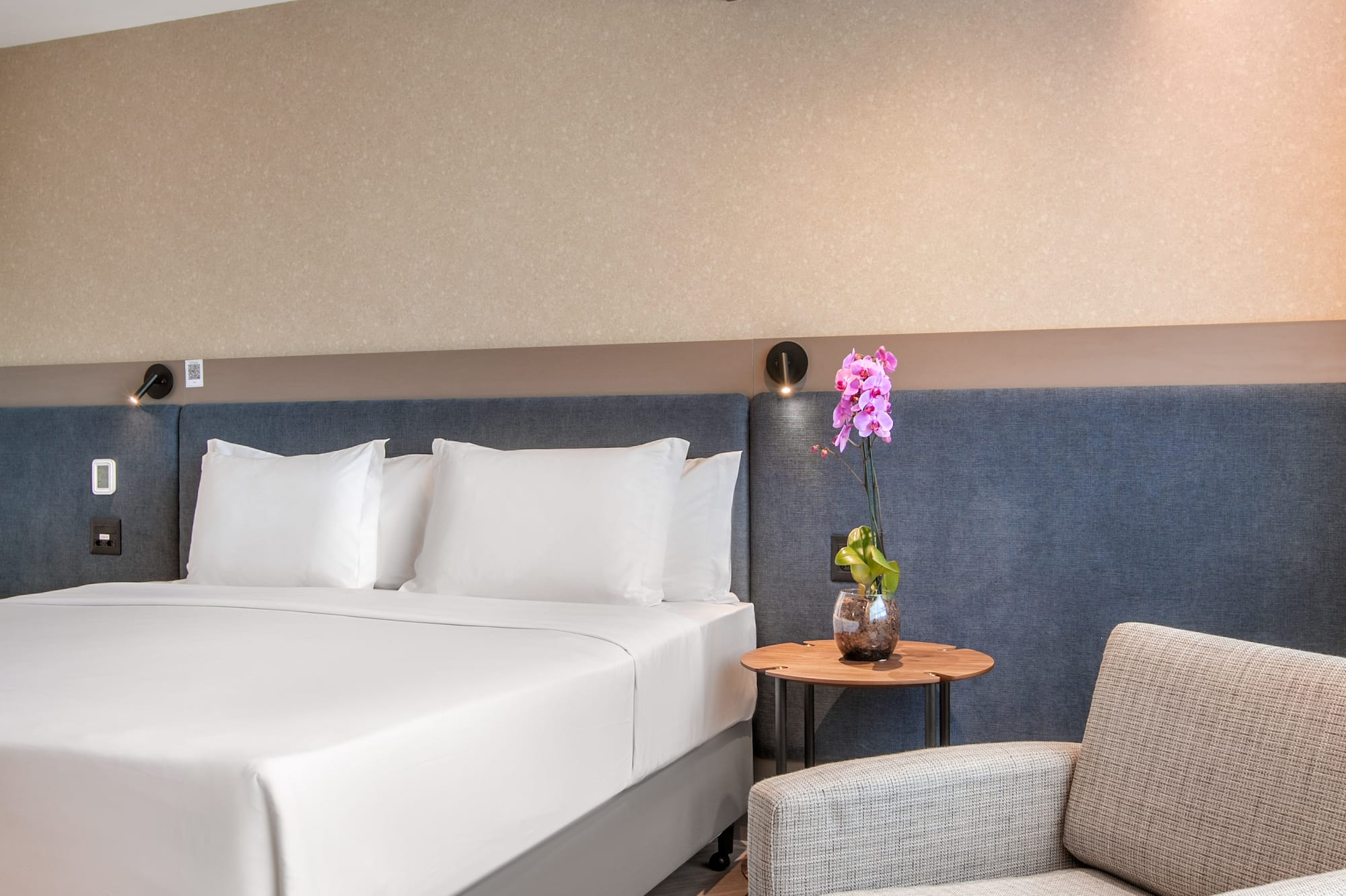 Contemporary hotel room with white bed, cozy armchair, and decorative orchid.