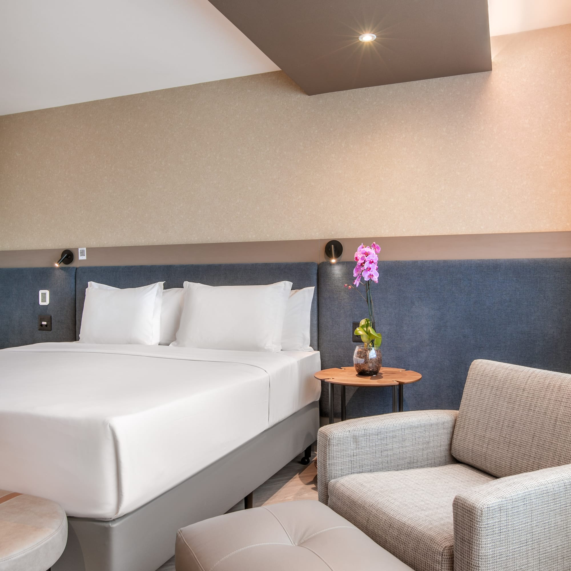 Modern hotel bedroom with white bed, gray armchair, ottoman, and orchid.