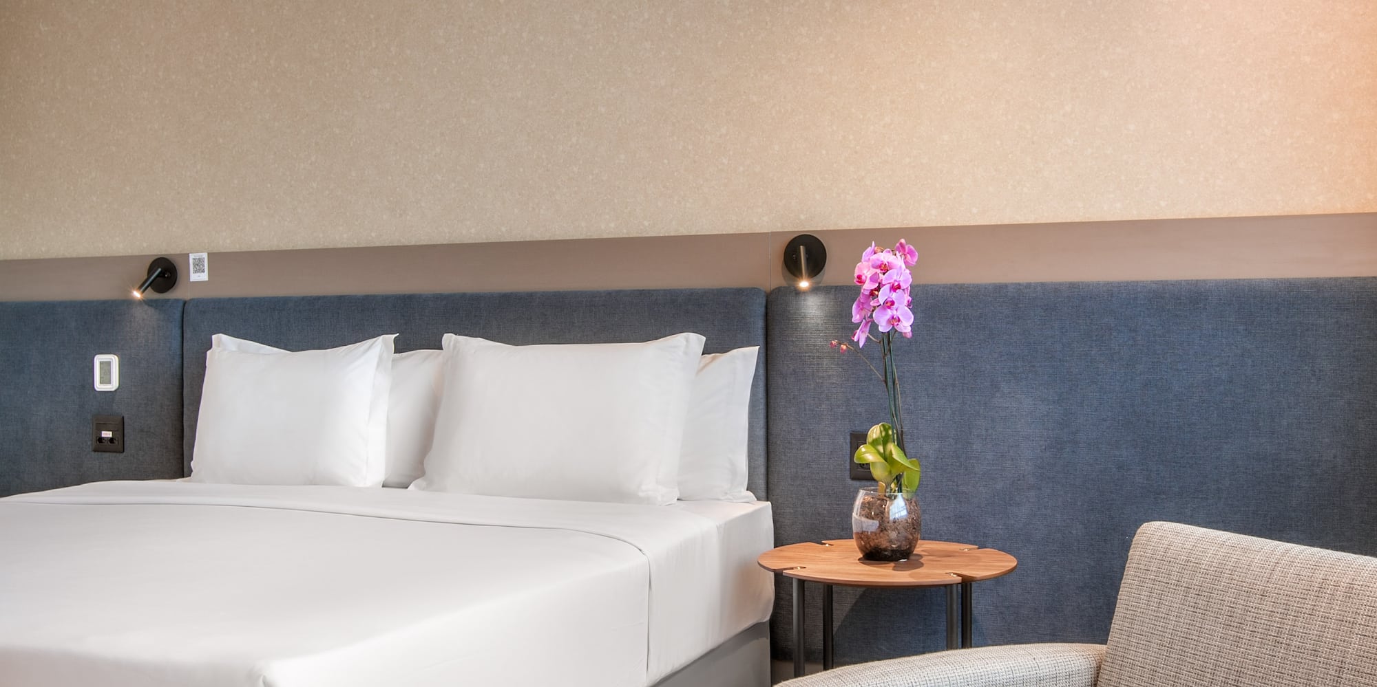 Modern hotel bedroom with white bed, gray armchair, ottoman, and orchid.