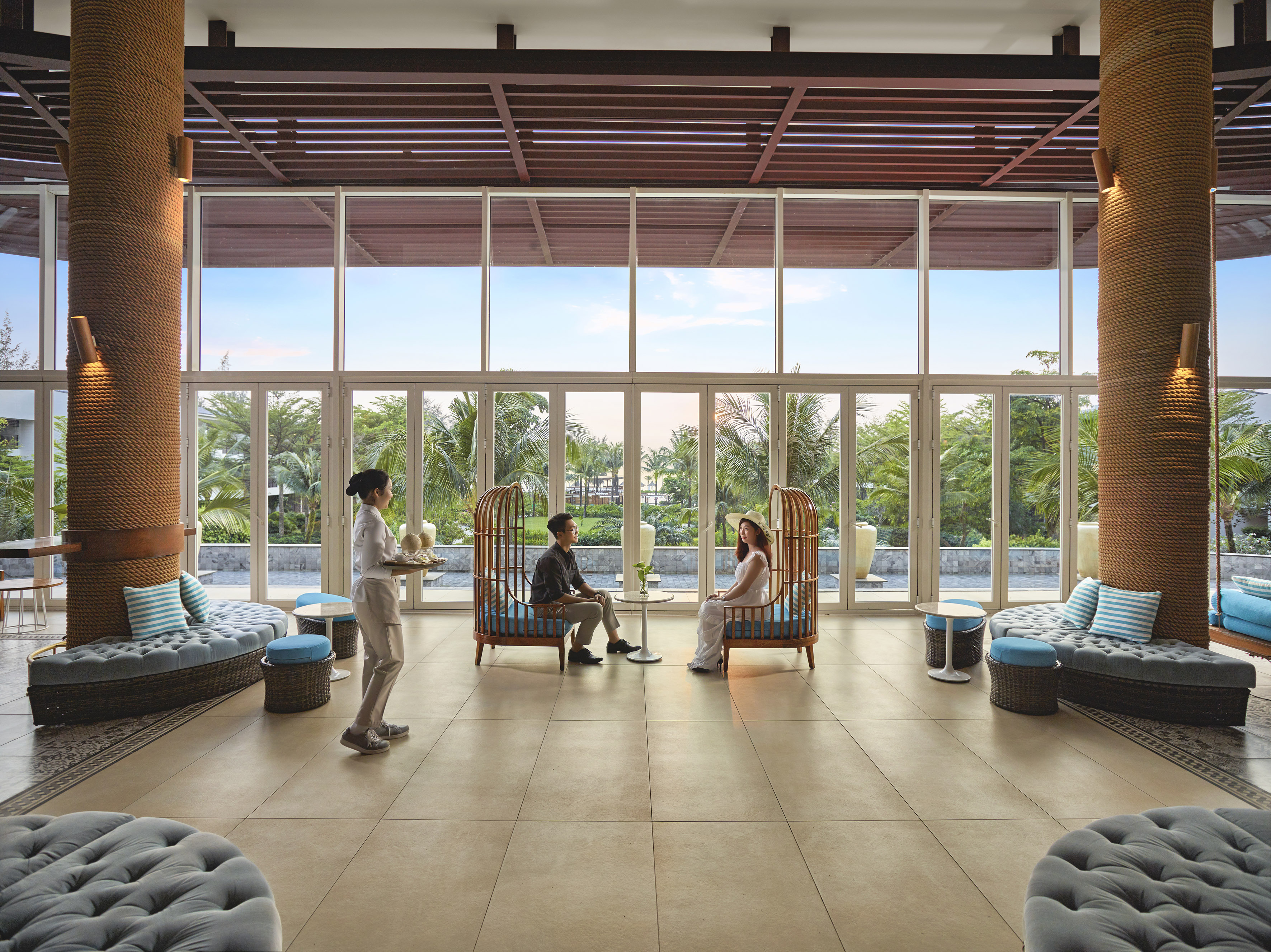 Bright resort lounge with couple on unique chairs, waitress, tropical view.